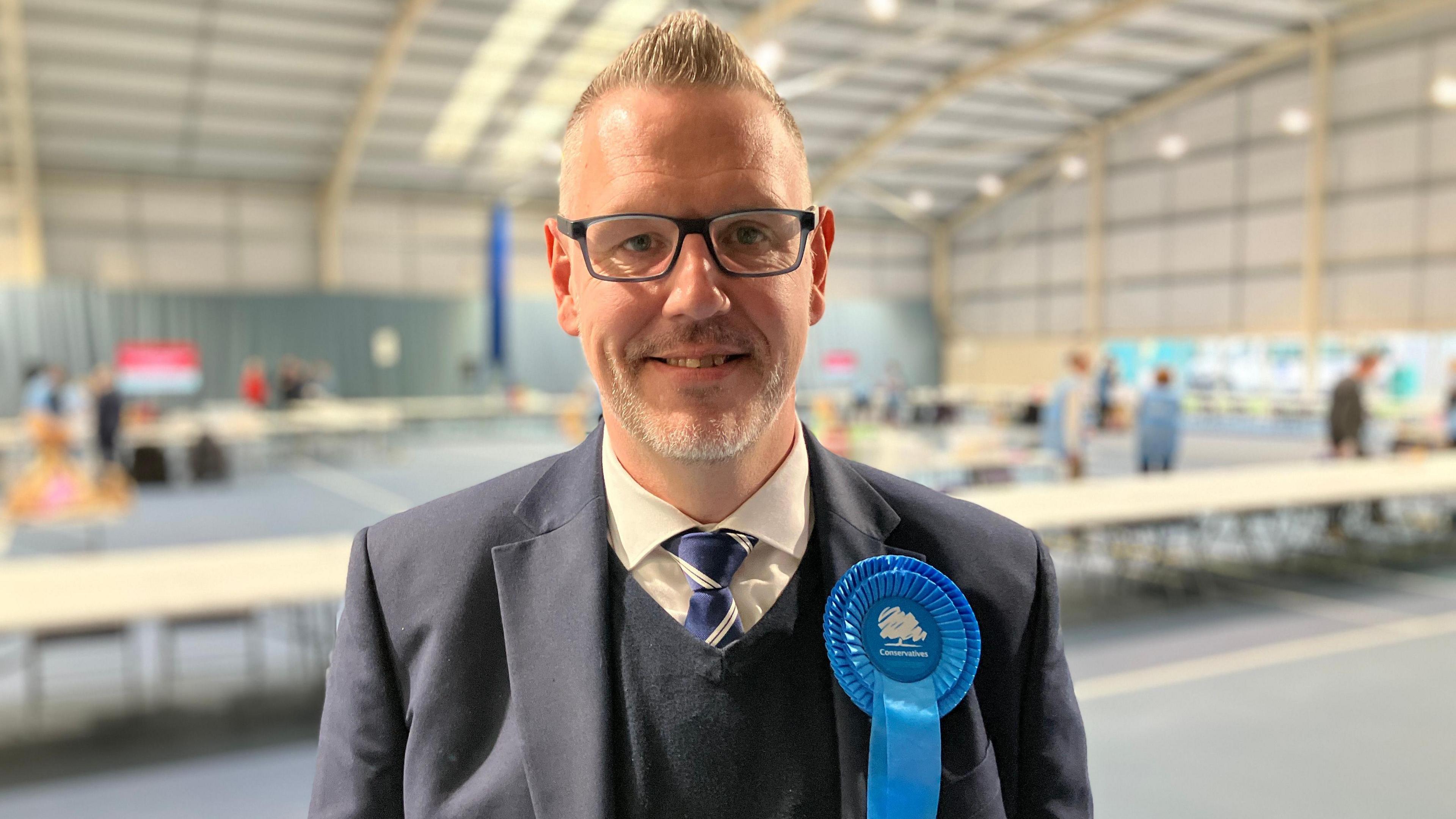 Mr Campion is pictured in an election hall - likely a sports hall or similar. The background is in a soft focus whilst Mr Campion is fully in focus - he's wearing a navy suit and tie and there's a blue Conservative rosette pinned to his left lapel. 