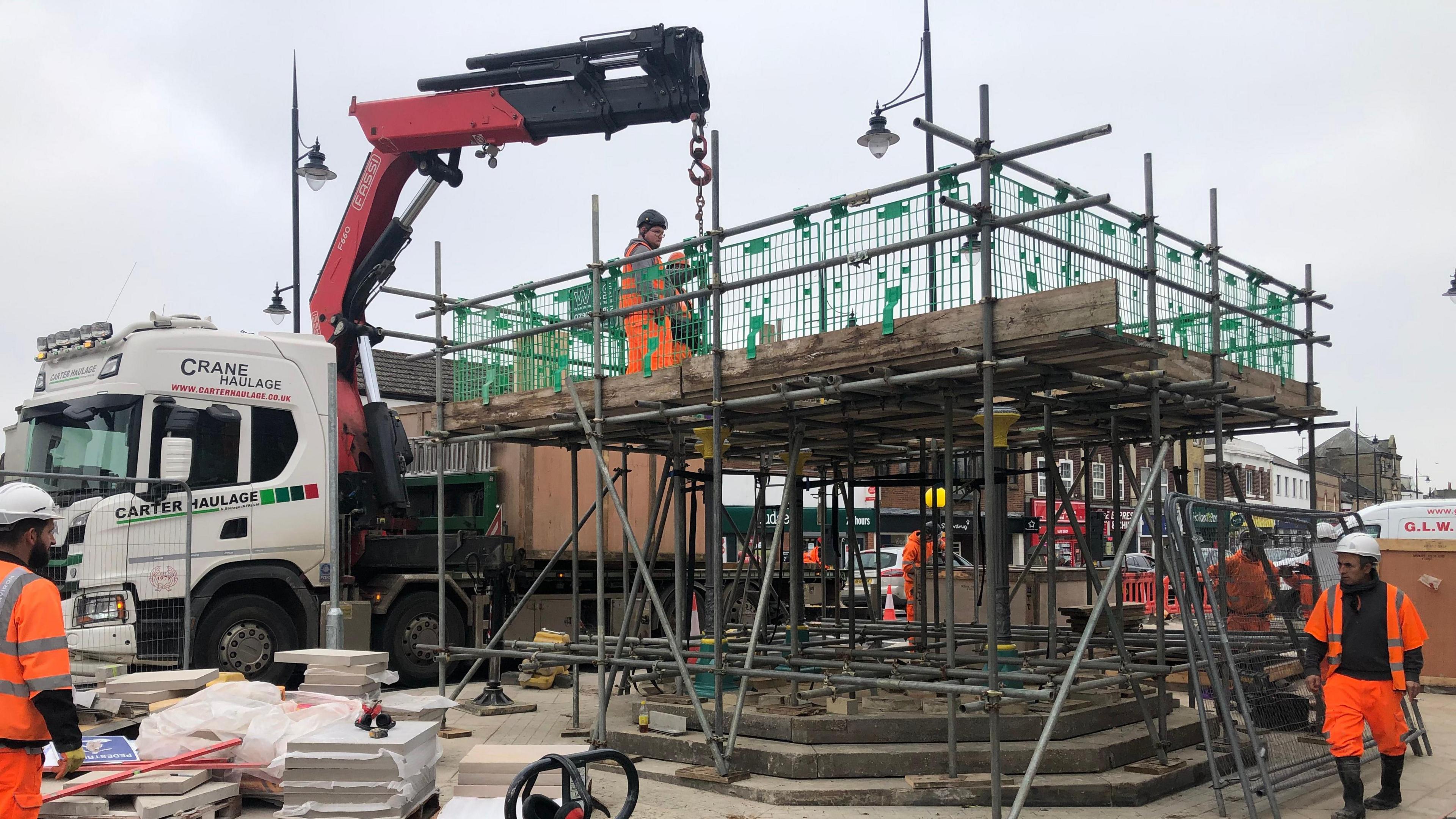 An orange and black crane arm is lifting sections of the fountain into place with scaffolding shrouding the site, there are three stone steps leading to the base of the structure and there are several workmen in high-viz orange outfits and hard hats on top of the 3m (nearly 9ft) high scaffolding and some below.