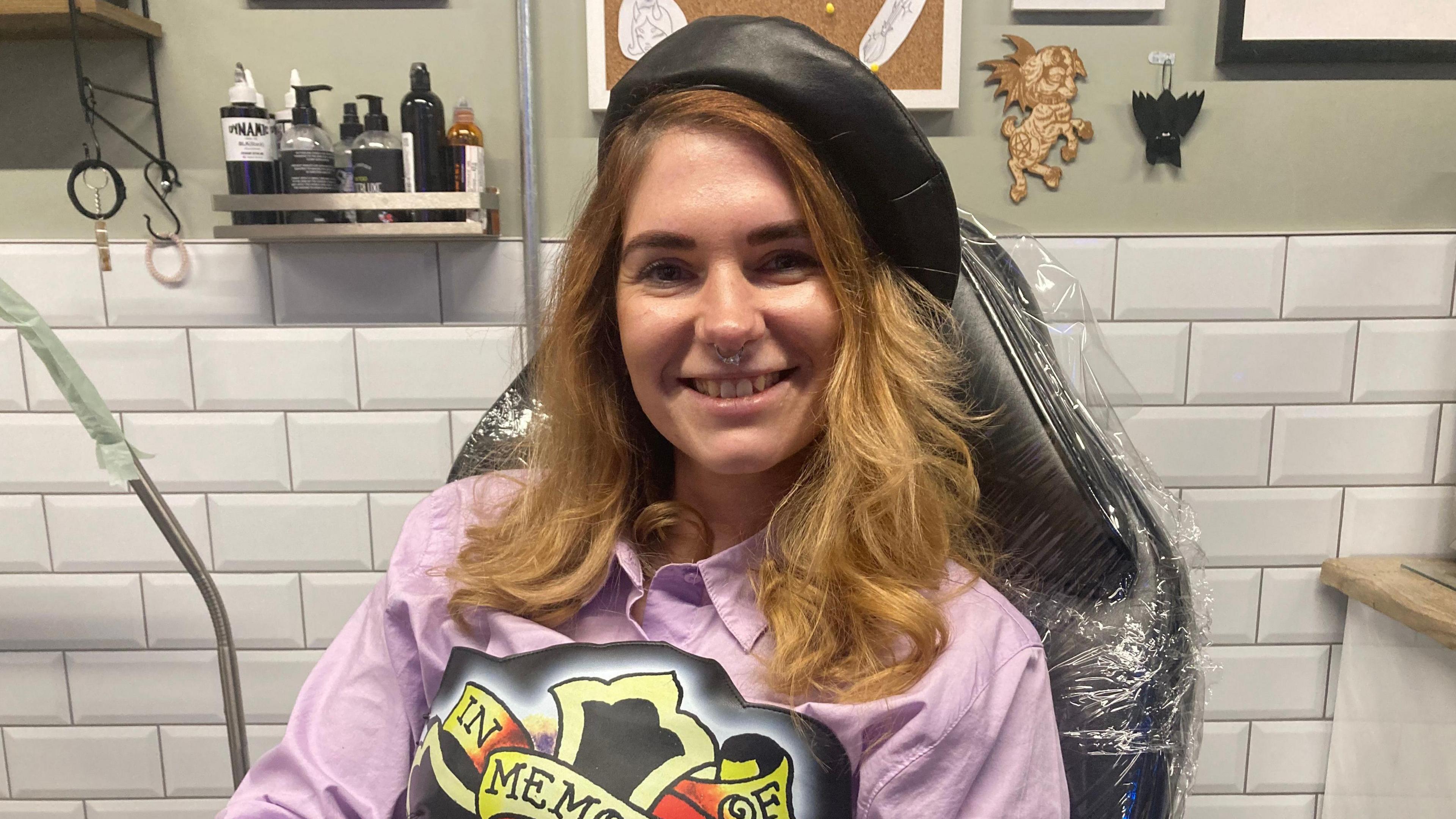 Alice Harding sits on a black chair covered in cling film smiling at the camera, she has light brown hair and a nose ring and is wearing a black beret