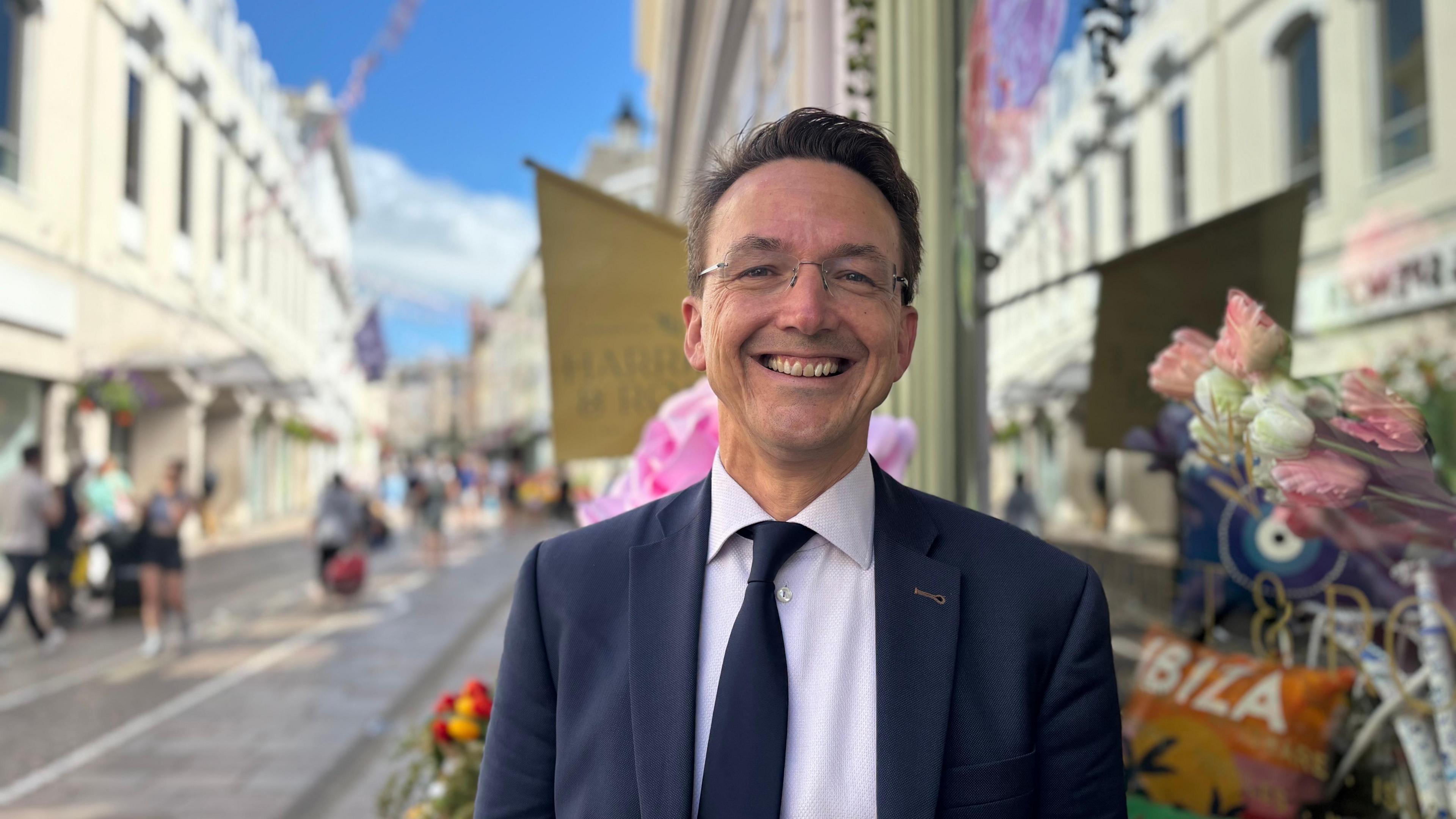 Deputy Kirsten Morel smiling at the camera. He is stood on the high-street and there is a shop to his left. In the reflection of the window, there are fake white and pink tulips.