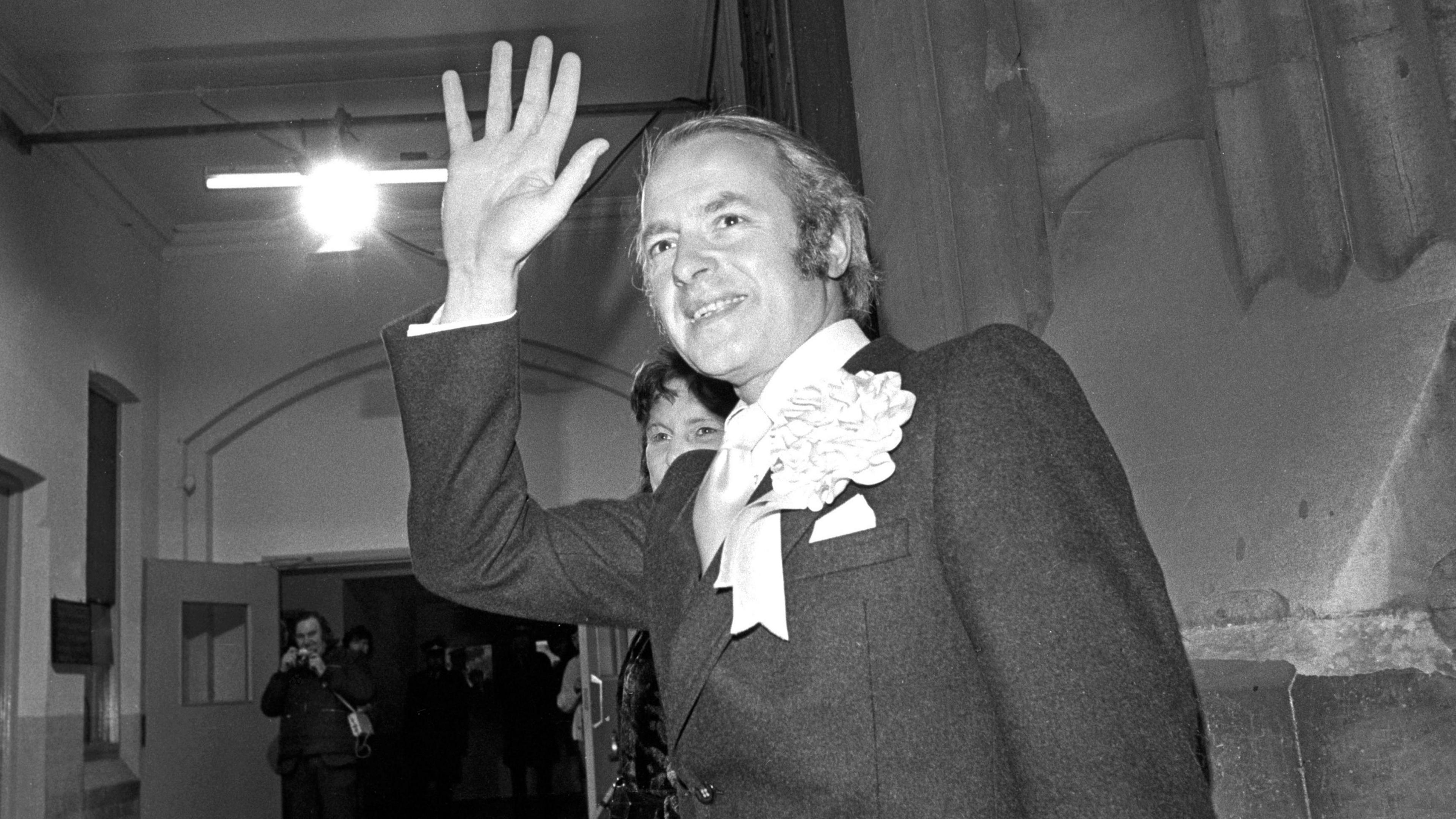 A black and white photograph of Dick Taverne with a rosette and matching tie.  He is waving at someone beyond the camera.