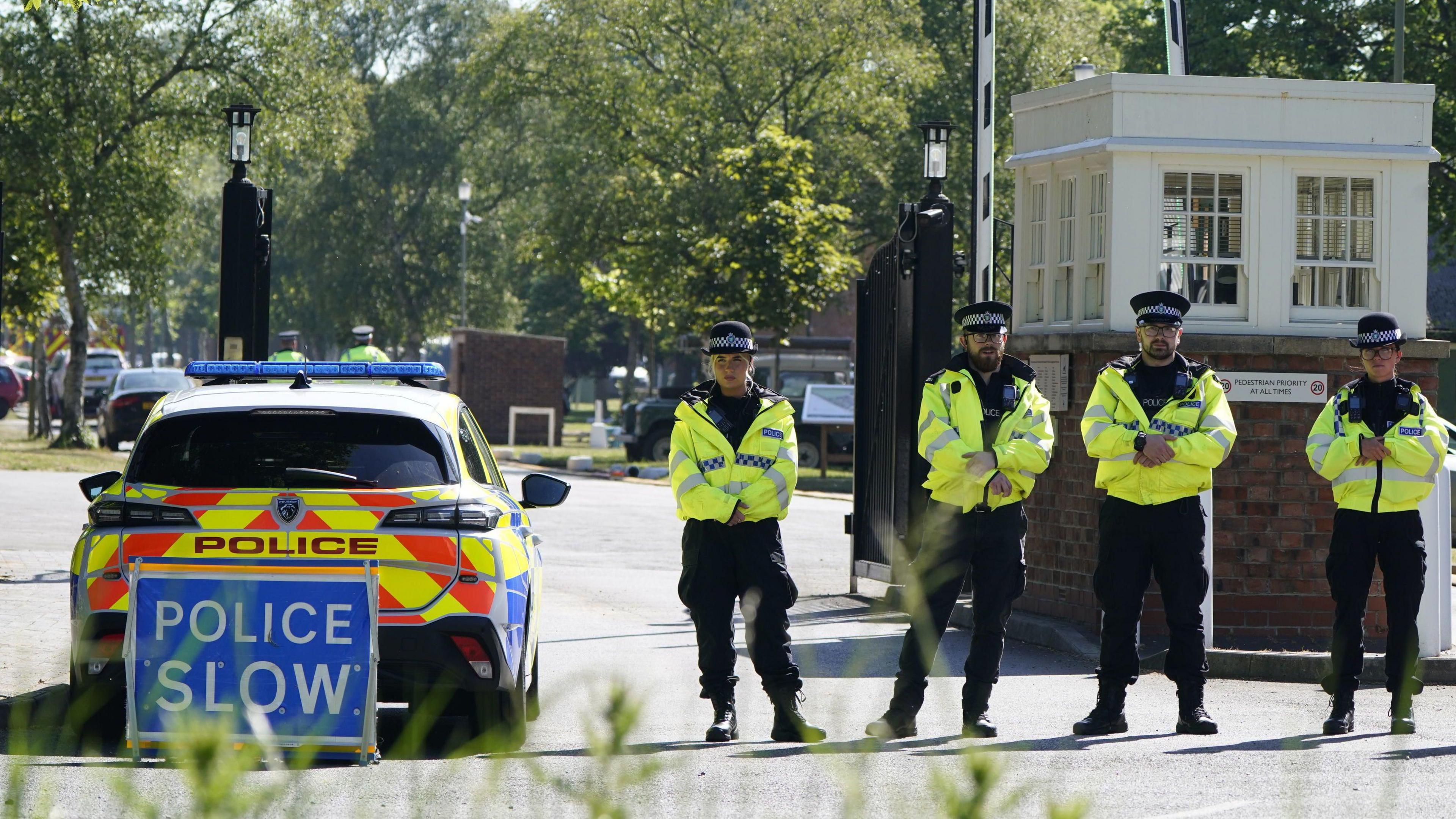 Police officers patrol at a cordon near the scene of the fire at Bicester Motion. Four officers with hi-vis jackets face the camera in a line on the right and there is a police car to the left. Behind the vehicle and facing the camera is a blue sign with white words that reads, "POLICE SLOW".