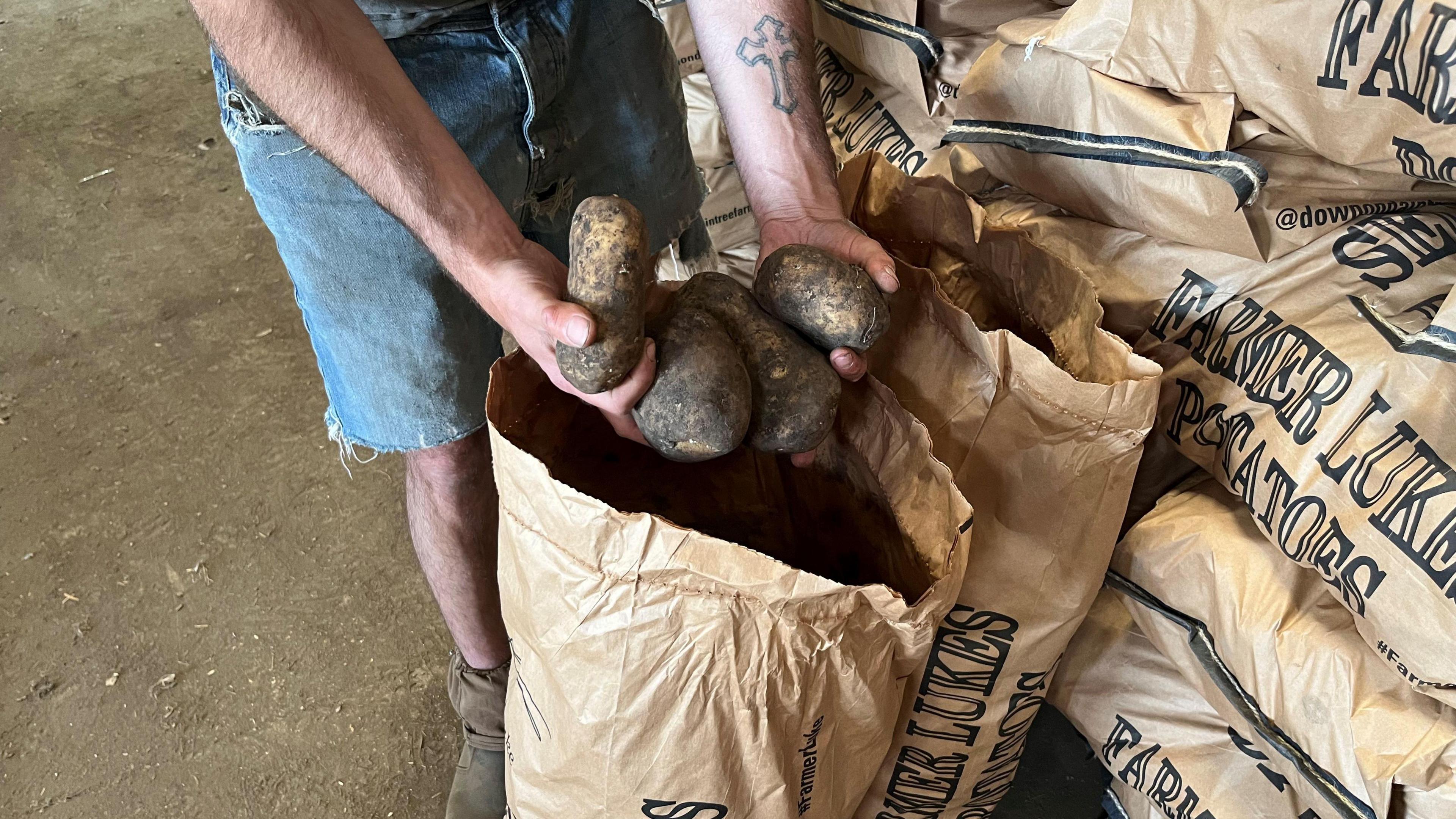 Mr Ablett's arms are visible as he shows off three large potatoes from an open brown paper sack, he has Demin shorts and brown ankle boots.