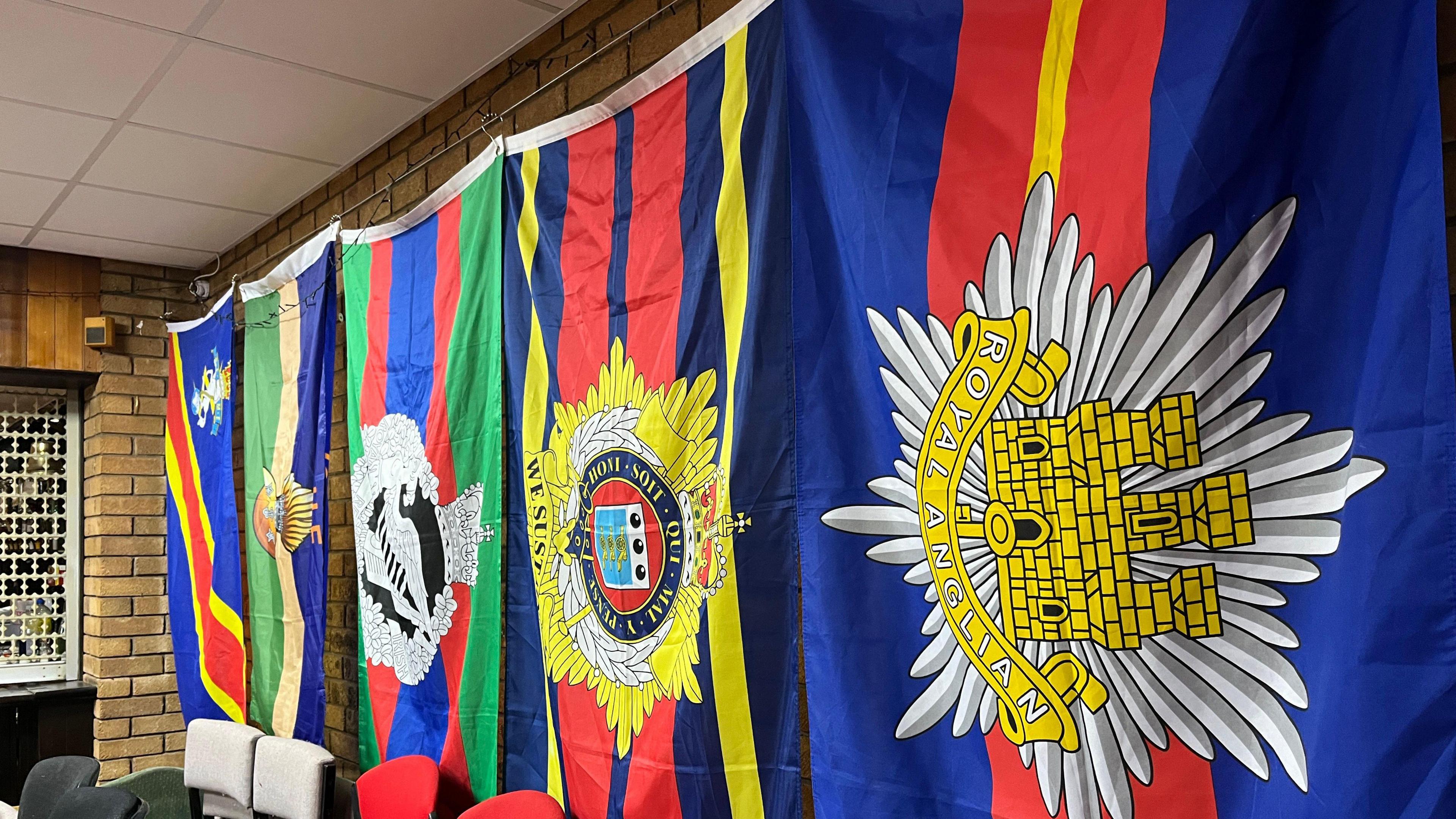 A row of brightly colour flags have been pinned against a brick wall inside the club . The closest to the camera reads 'Royal Anglian'. 