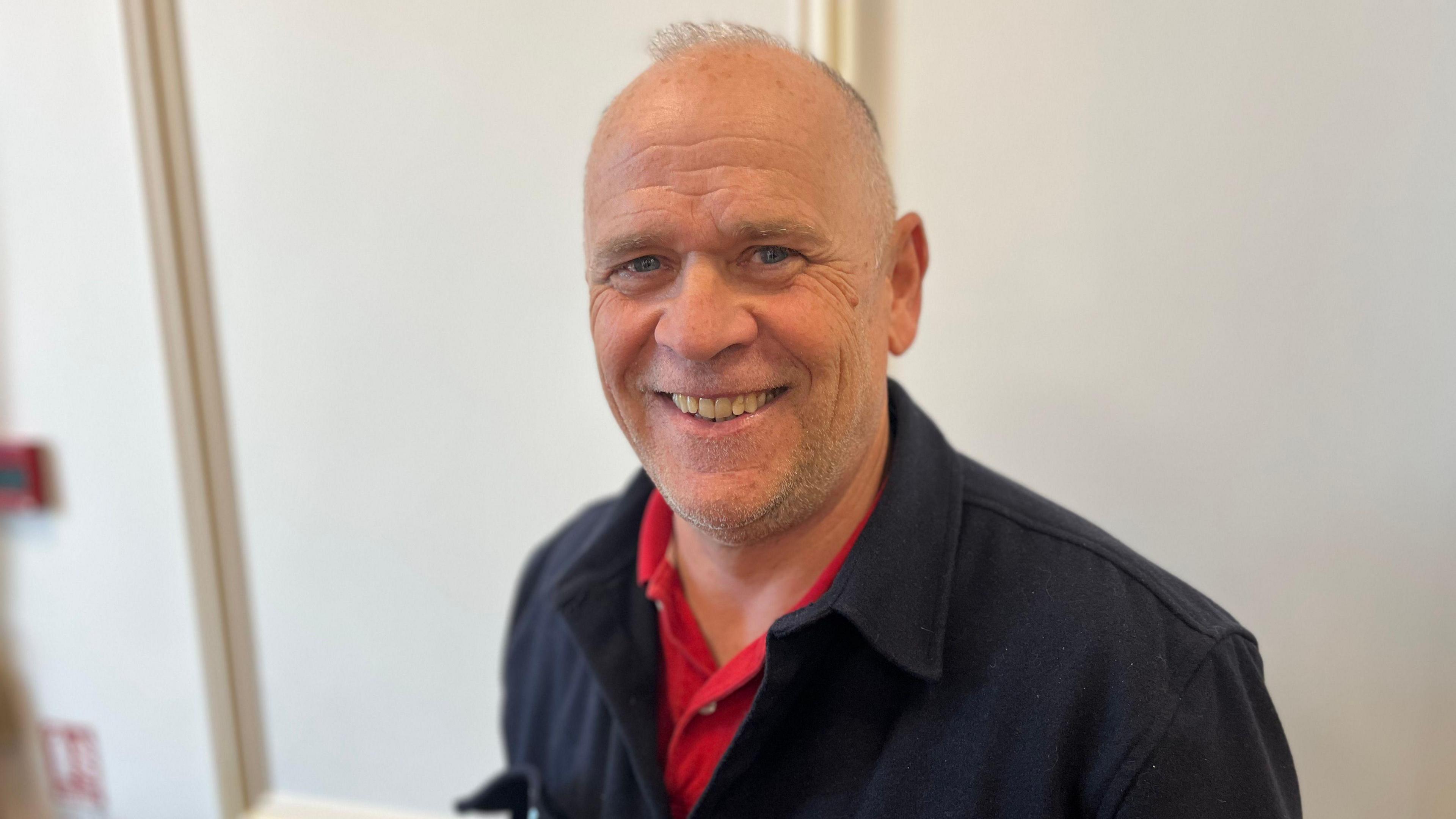 Marc Laine smiling and looking at the camera. He's wearing a red shirt and a navy jumper.
