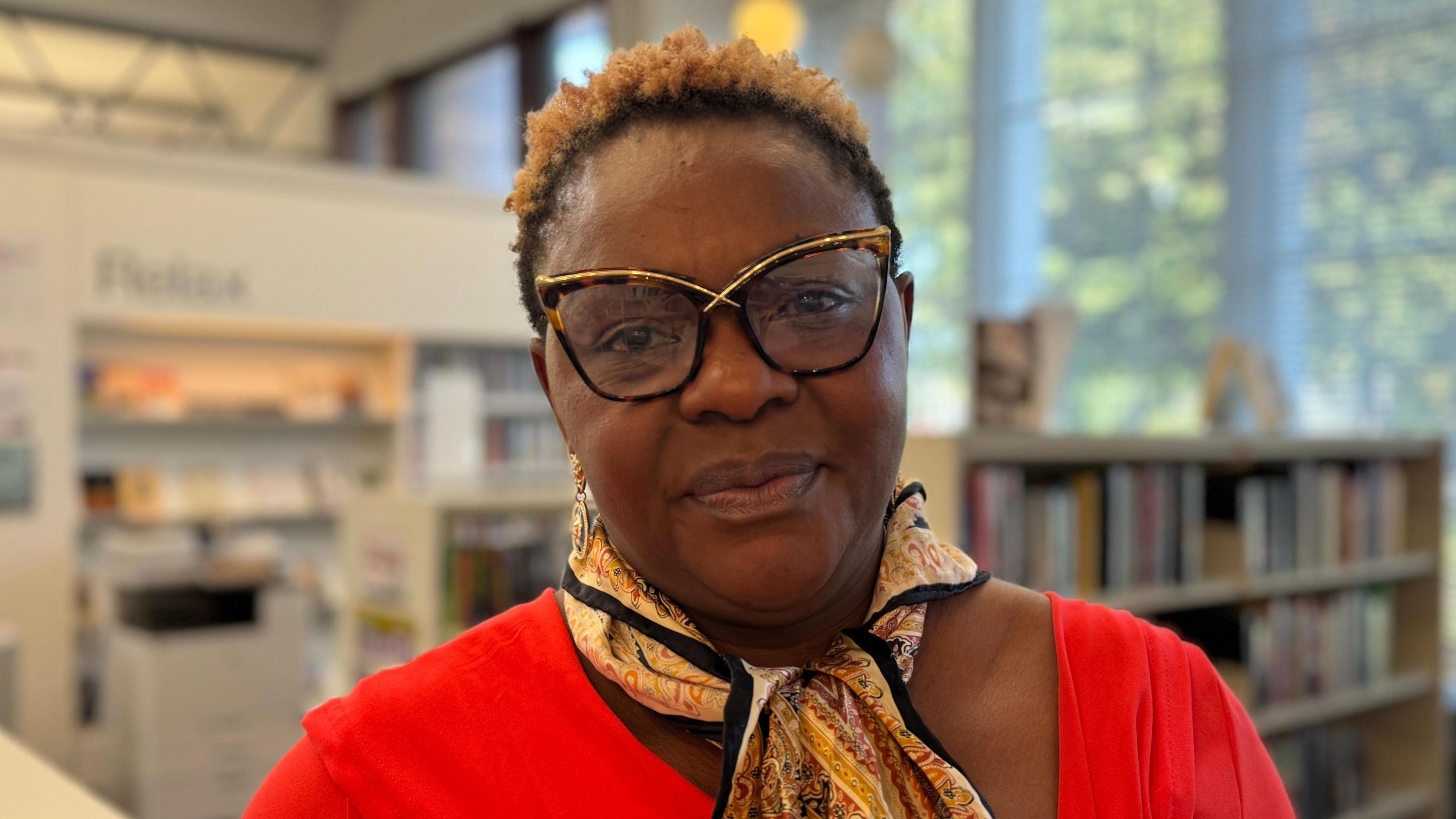 Alexis Ekwueme - A woman with short hair wearing gold and tortoise shell rimmed glasses, a patterned scarf and a red top looks at the camera. She is standing in the middle of Ormeau Road library.