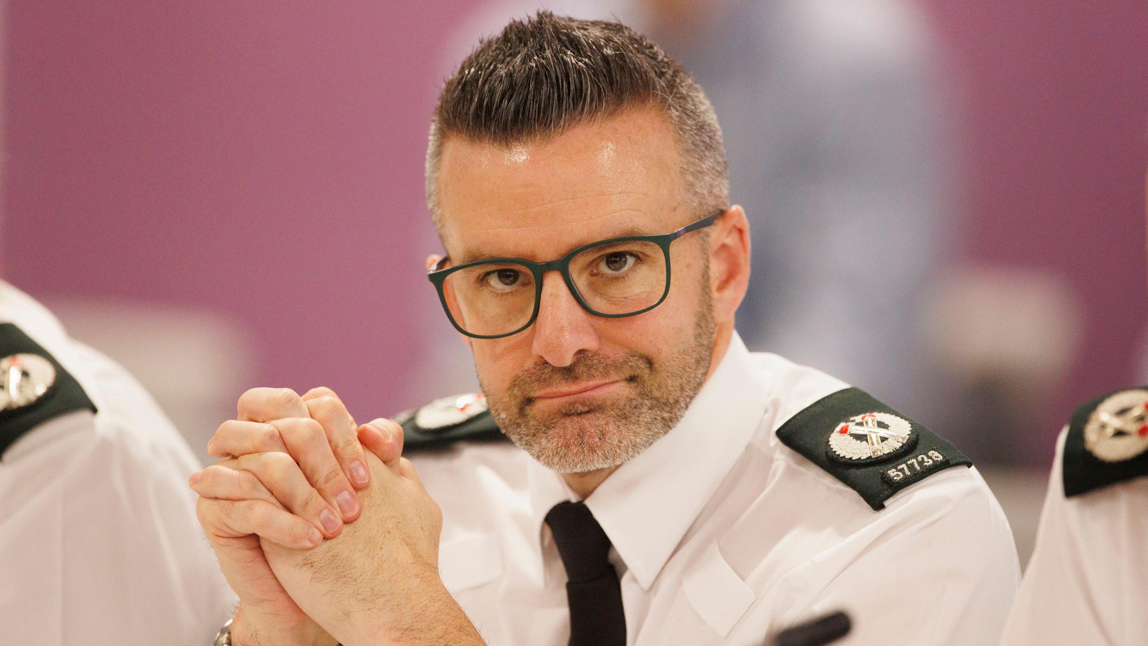 Bobby Singleton - a man with short, dark and grey hair wears square framed glasses and is wearing a police uniform. It includes a white, collared shirt, black tie and embroidered shoulder lapels.