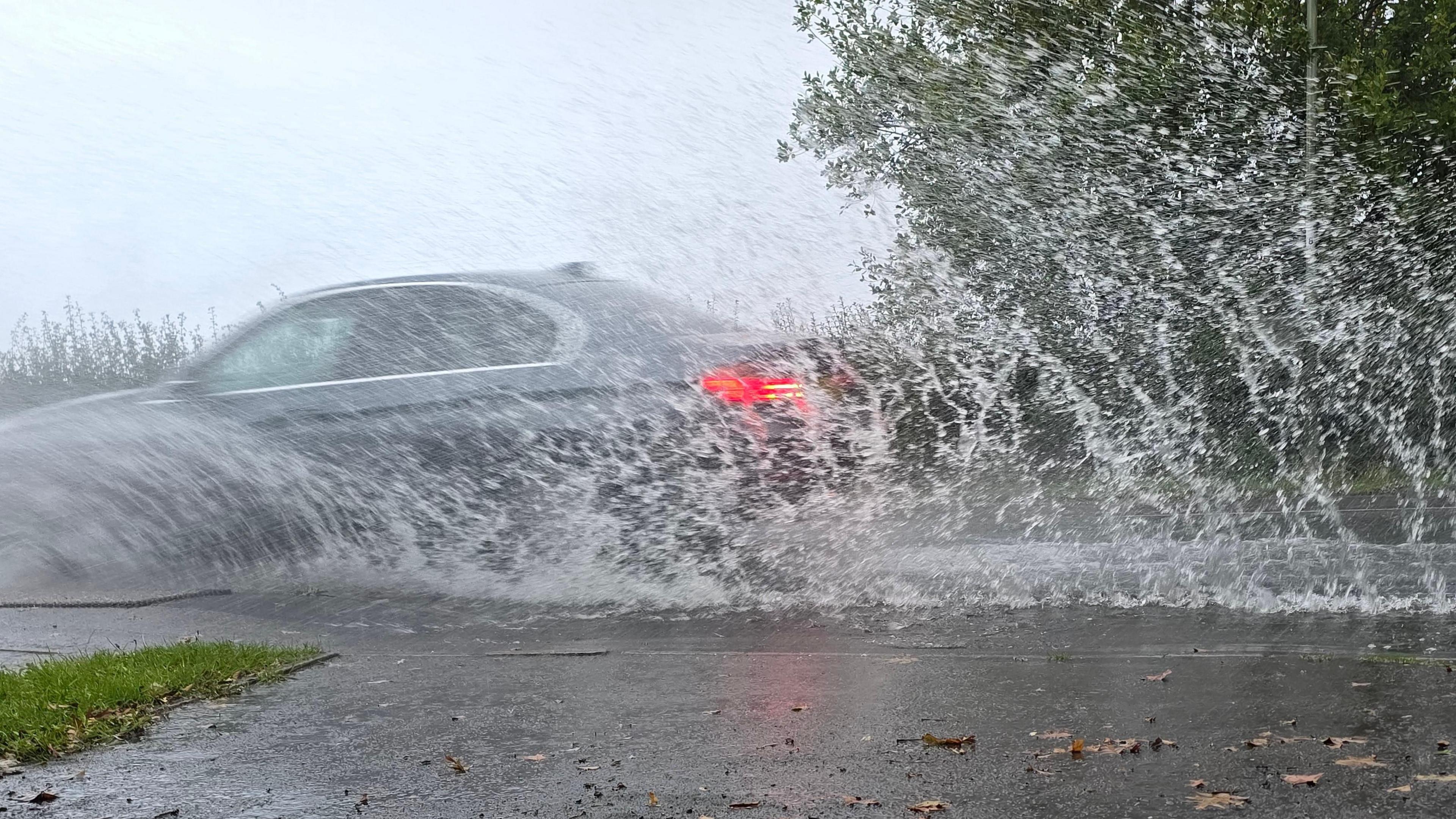 A car drives through a roadside puddle throwing up a curtain of water in its wake. The cars rear red lights can be seen. The pavement in the foreground is wet with some fallen leaves on it and a tree stands in the background. 