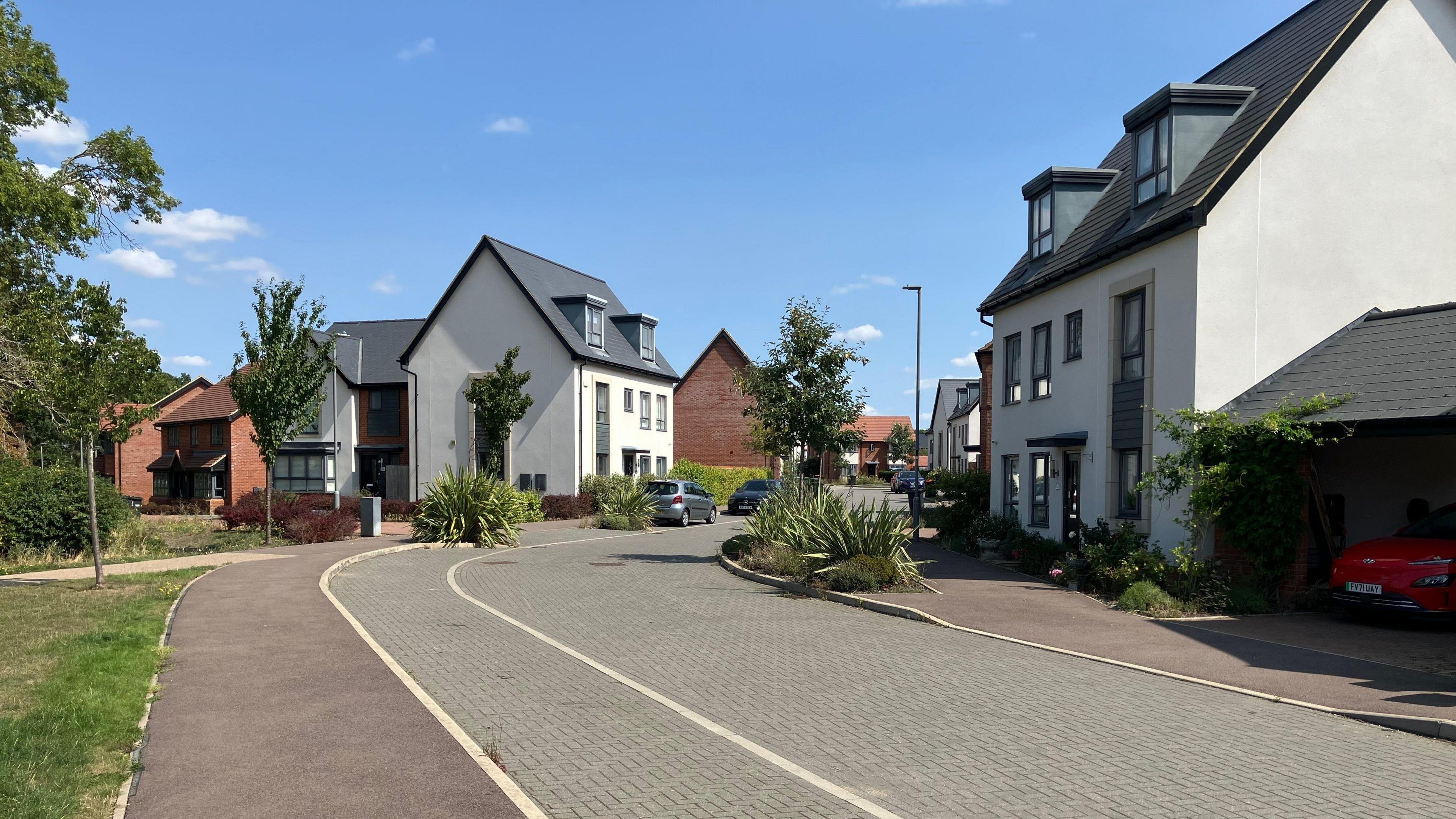 A street on a modern housing estate. To the right of the picture is a large white house with a slate roof and a slate covered parking area to the side where a red car is parked. In the foreground to the left of the picture is a grass verge and pavement with a road going down the centre of the picture. In the background a number of white painted and red brick houses can be seen.