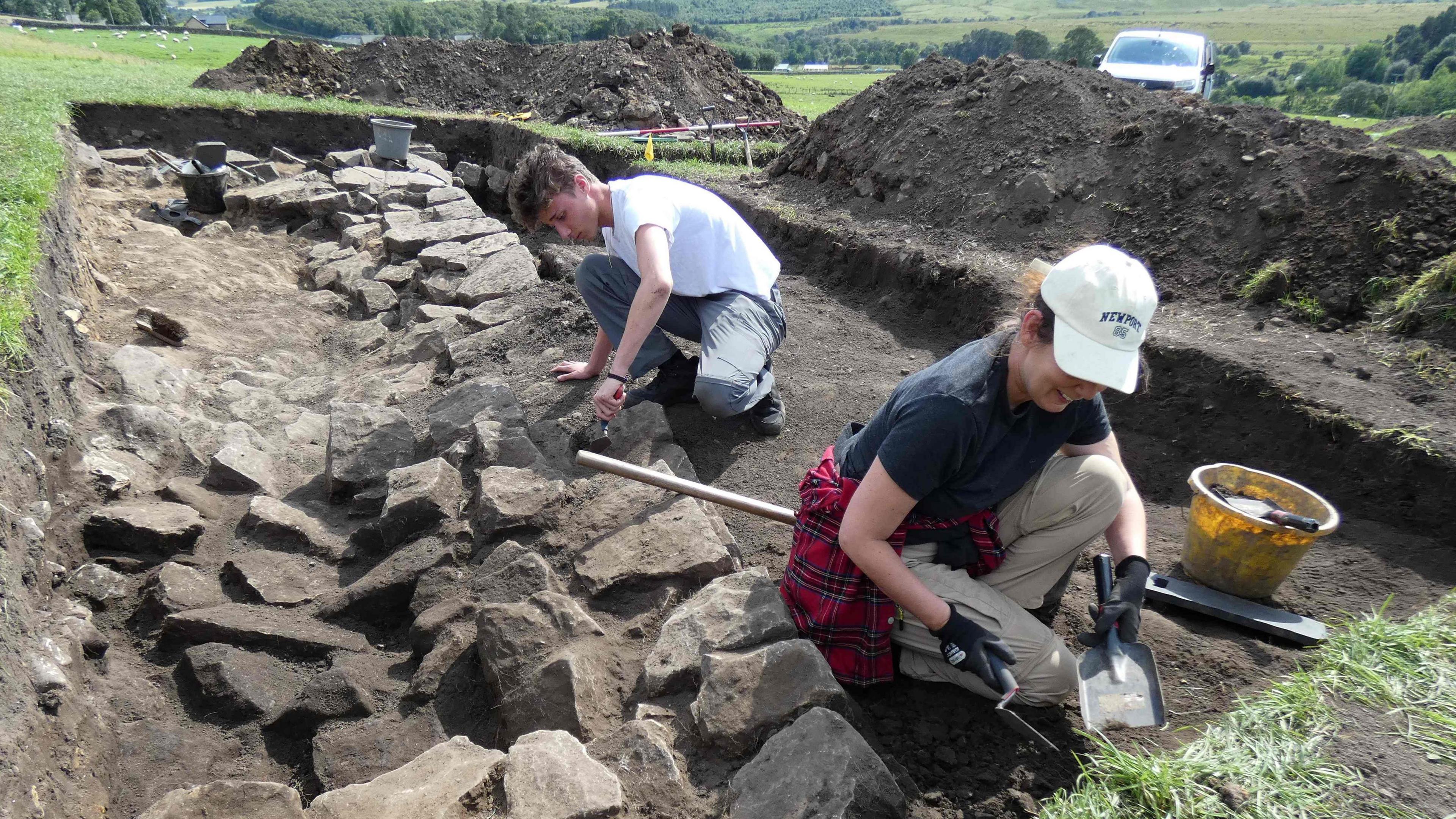 Two volunteers are carefully using trowels to explore a trench dug next to an old Roman wall. Heaps of dirt can be seen piled next to the trench.
