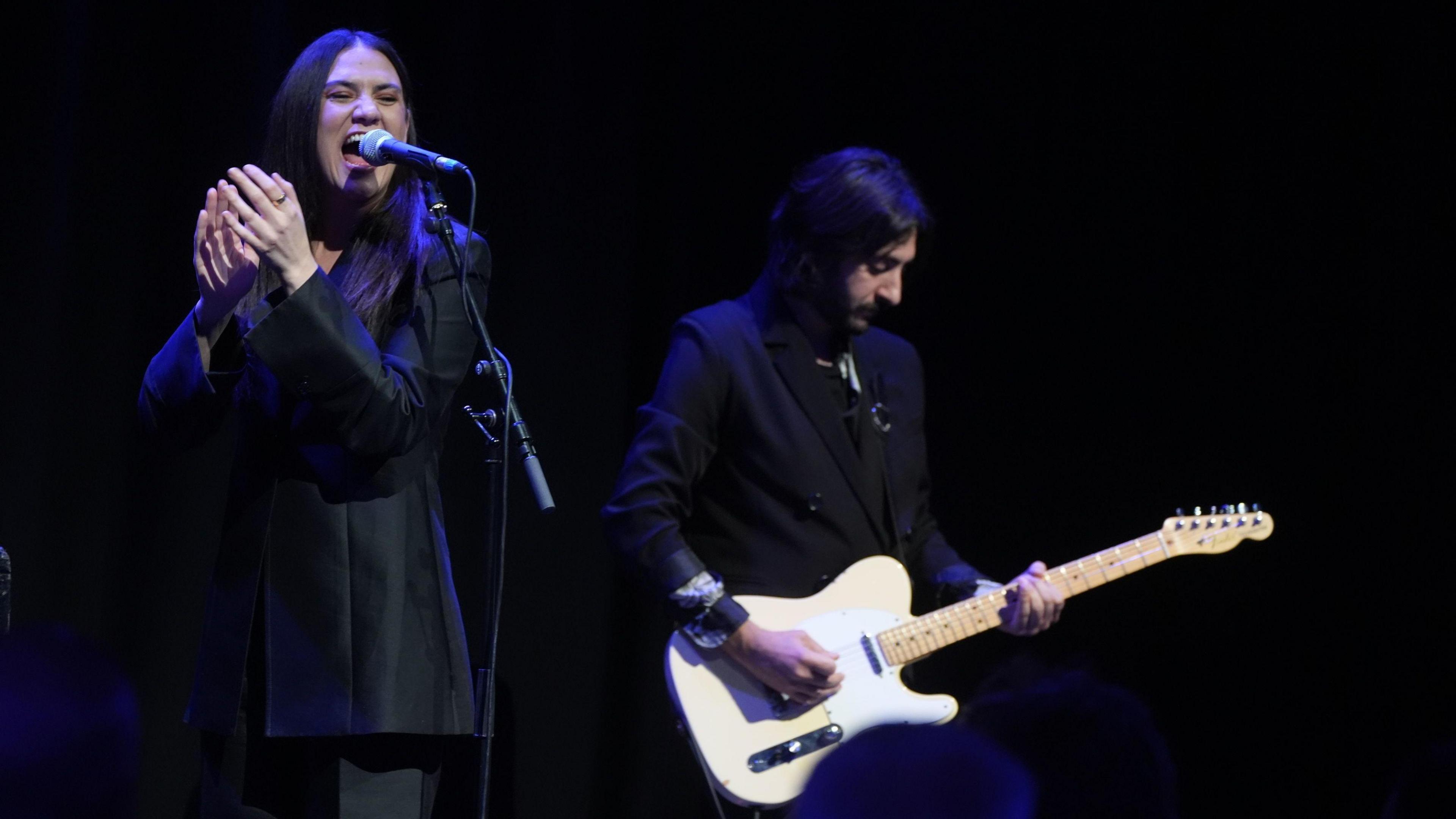 Nadine Shah sings into a microphone alongside a man playing a guitar. She has long black hair and is wearing a black suit. The guitarist also has black hair and a black suit. His guitar is painted cream.