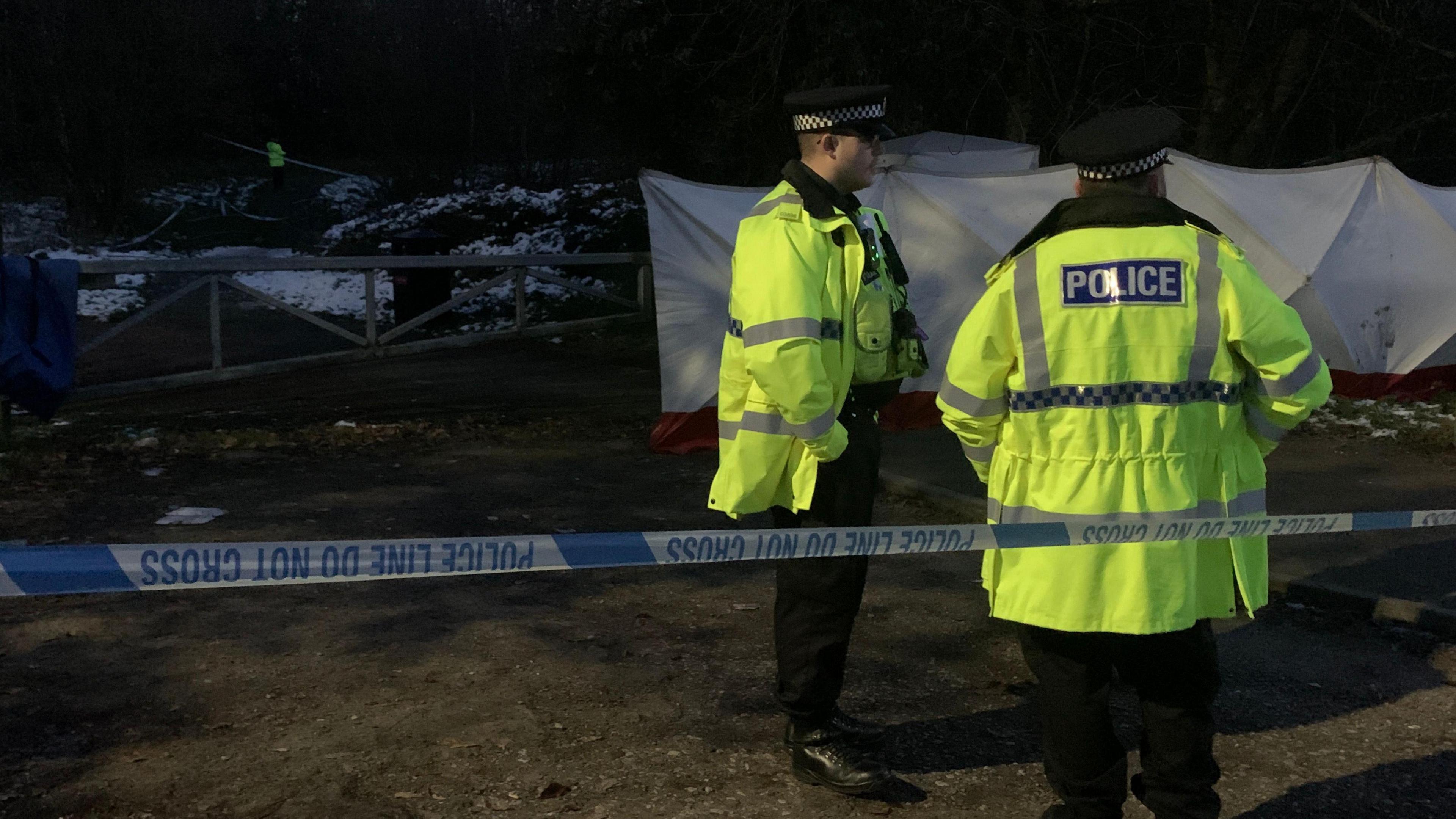 Two male police officers in reflective yellow jackets stand in a paved area cordoned off with police tape. In the background is a white forensic tent and a wooded are dusted with snow