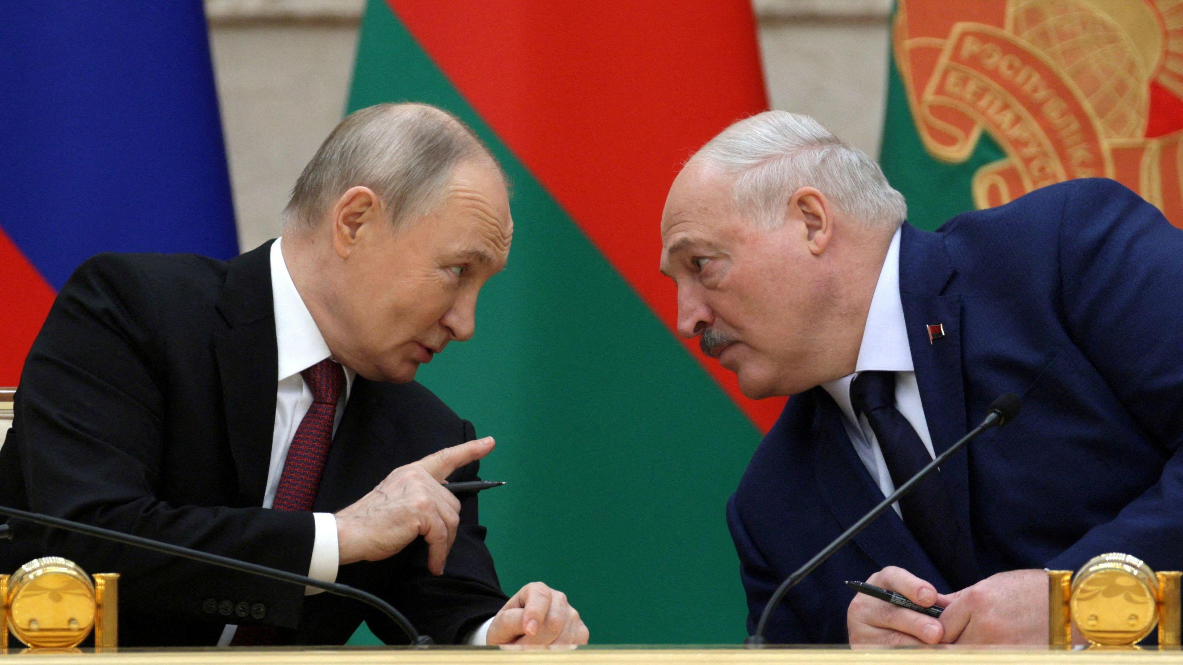 Presidents Vladimir Putin and Alexander Lukashenko meet at a state council. The Belarussian and Russian flags are behind them.