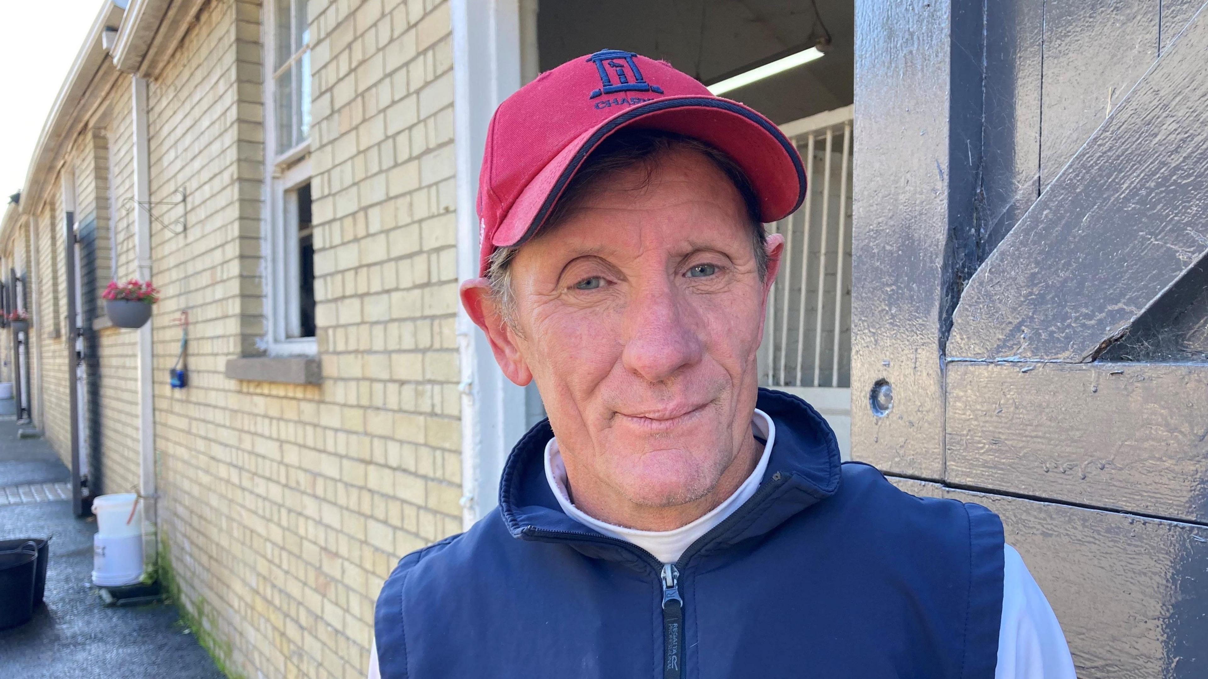 Joe Scally, wearing a red/pink baseball cap and blue gilet looks directly at the camera. He has blue eyes and is standing in front of a stable door.