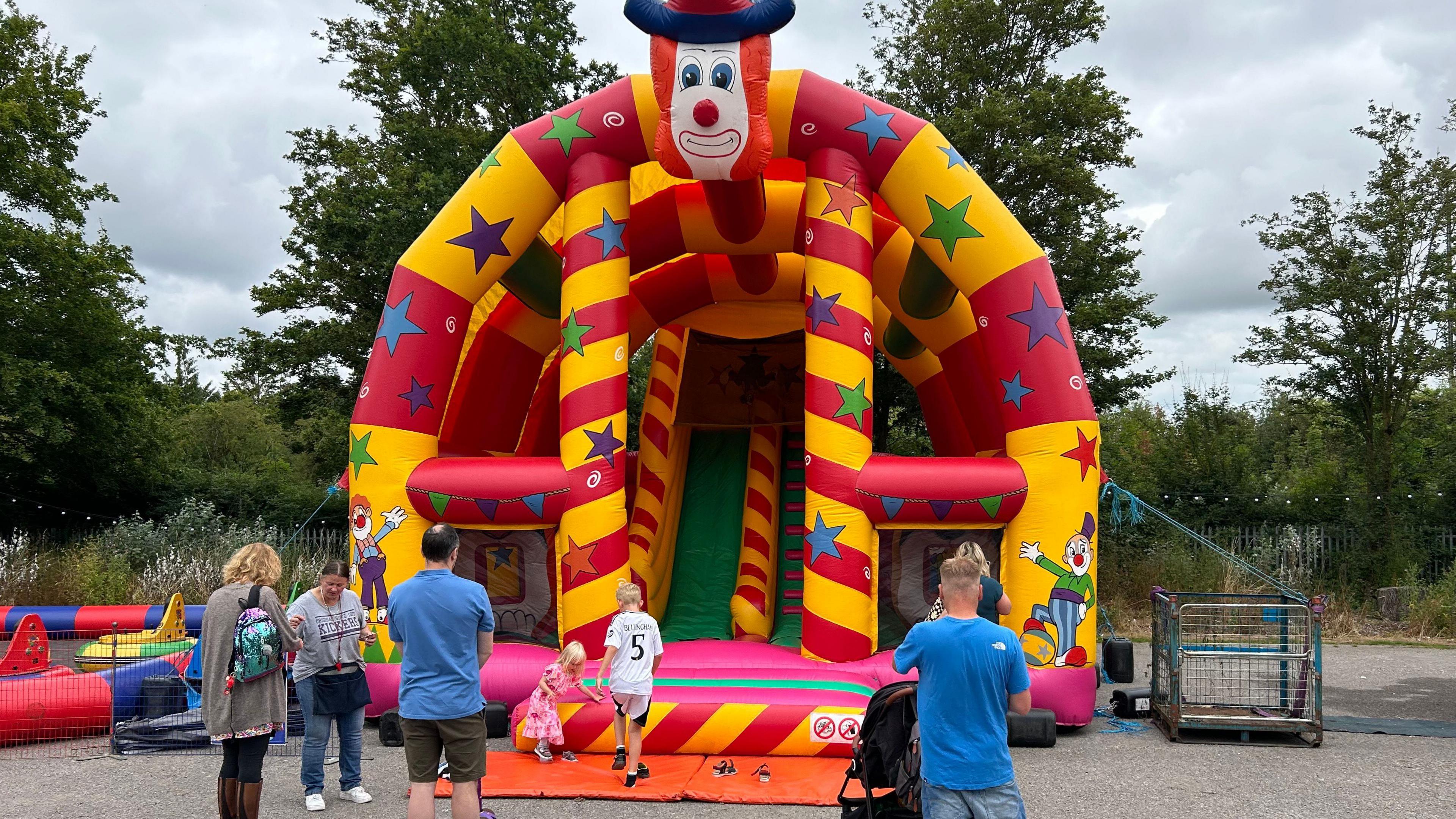 Inflatable bouncy castle and slide. It is multicoloured covered in stripes and stars and has a large clown head at the top. Children and parents can be seen near it.