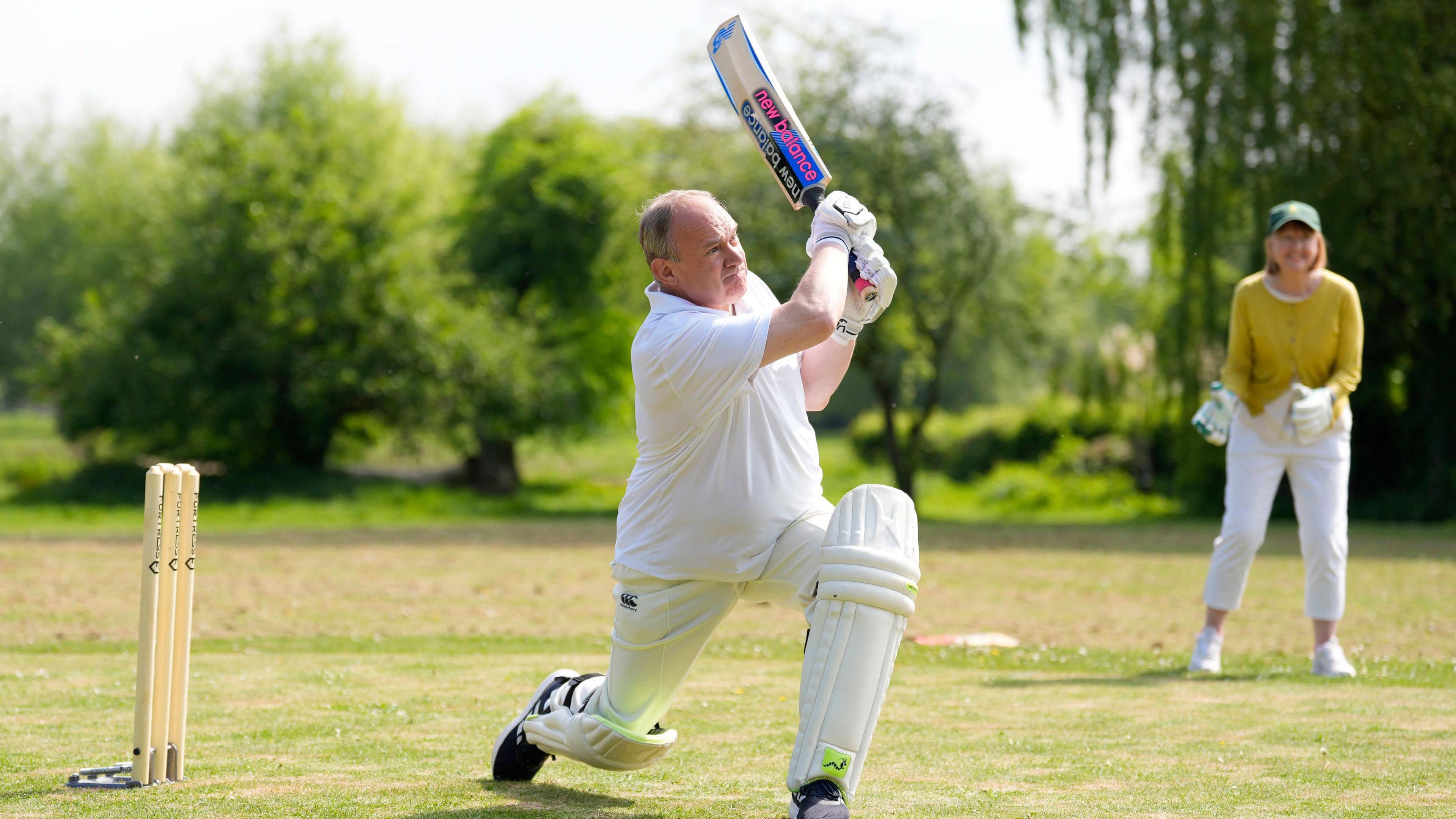 Sir Ed Davey lunges forward as he swings his cricket bat. A woman is in the background of the photo smiling and waiting to catch a ball.