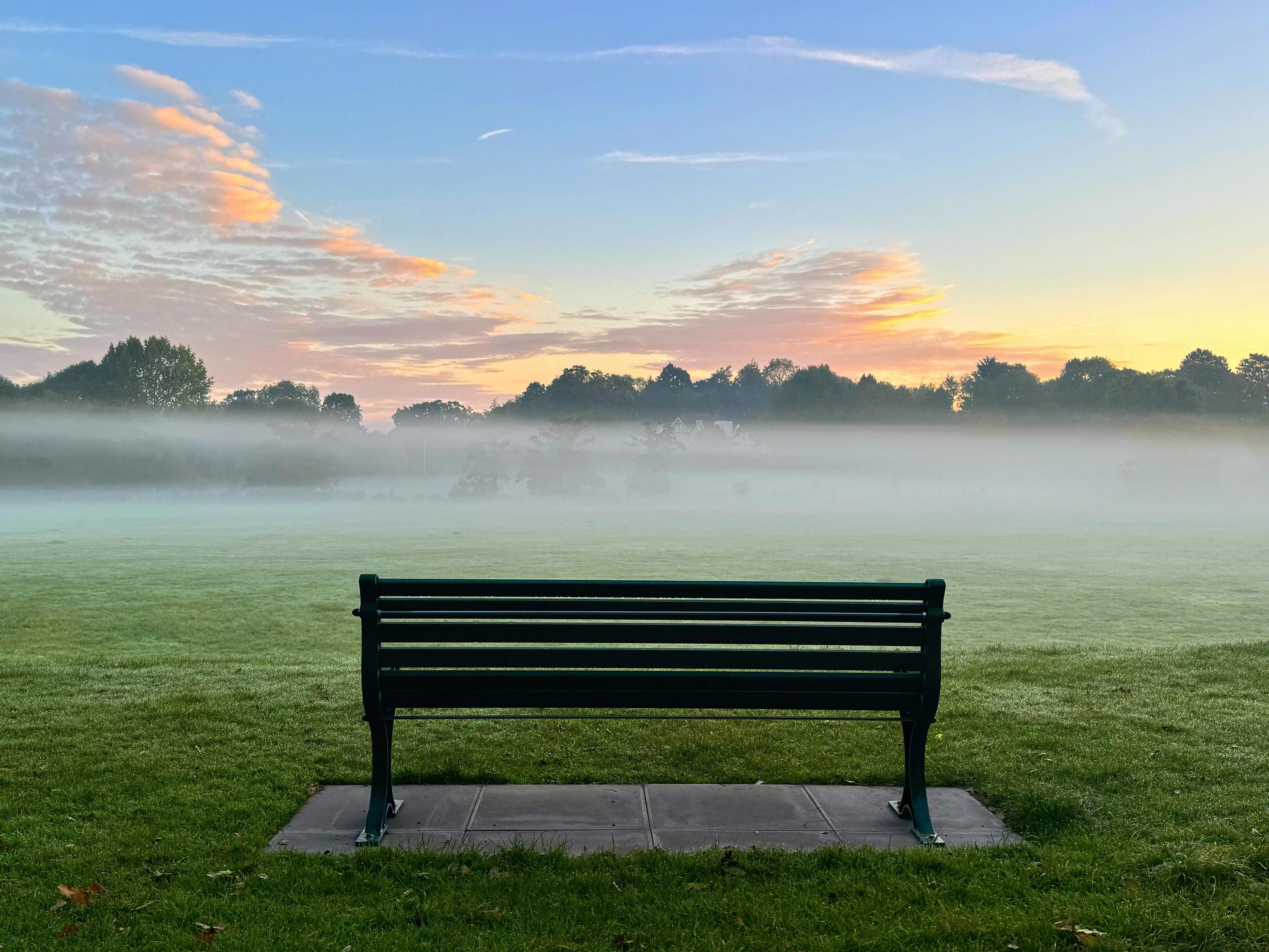 A clearly defined layer of mist lies over grassland below a blue sky with the sun starting to rise behind trees. In the foreground, a traditional park bench sits on paving slabs.  