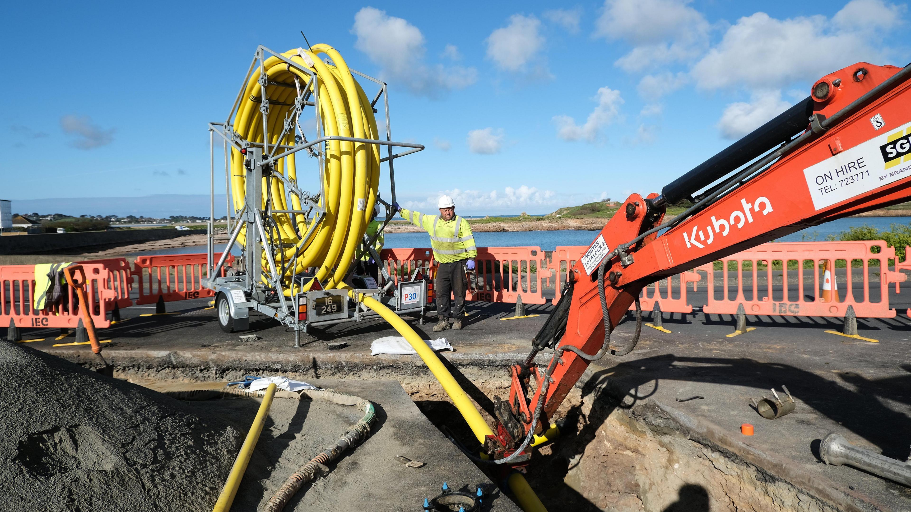 An orange hydraulic arm fits yellow cabling into a trench dug in the road while a workman in high viz clothing and a white hard hat supervises.