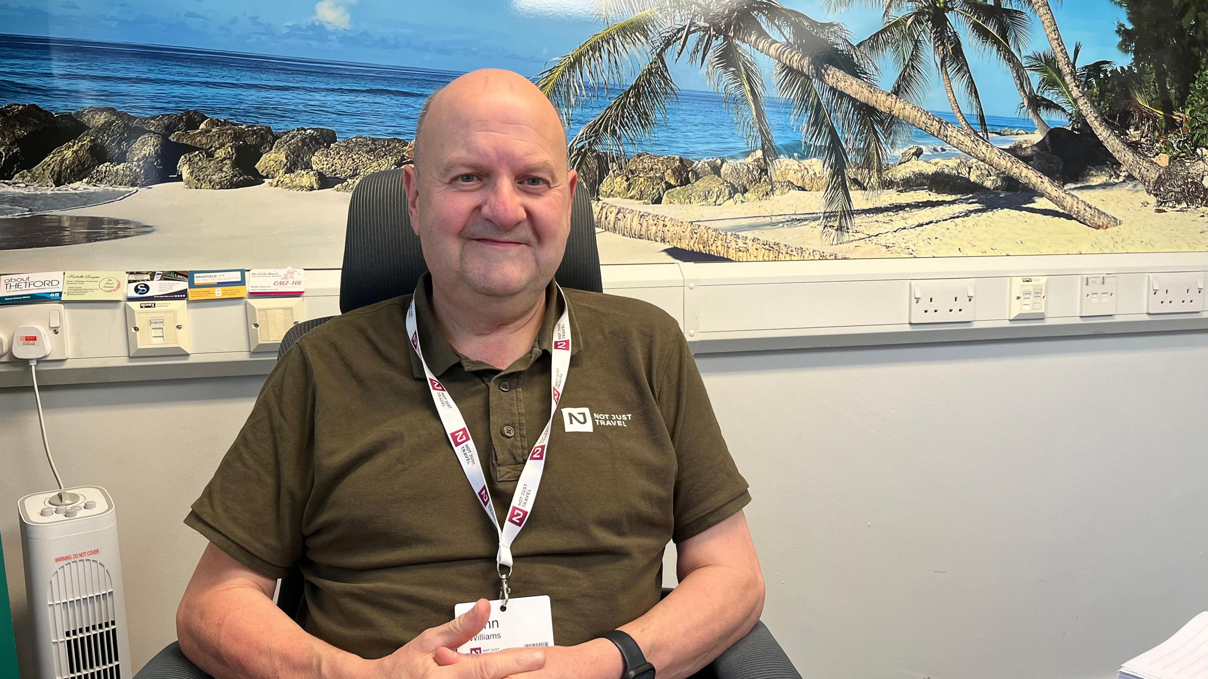 John Williams runs NotJustTravel at the Charles Burrell Centre. He is wearing a olive green branded polo shirt. Behind him is a picture of a beautiful white sandy beach with bright blue sky and sea.