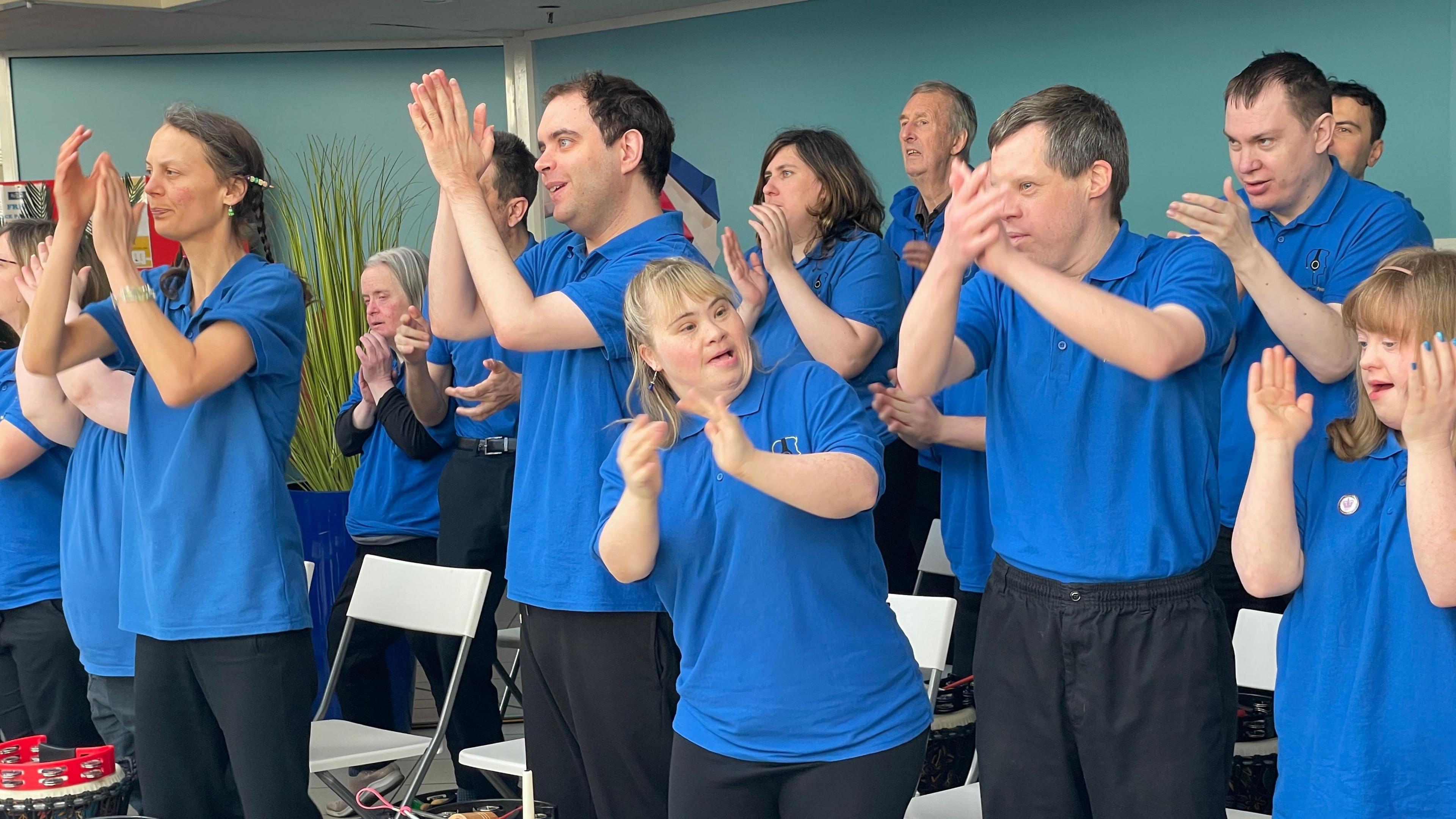 Singers from the Music Man Project wearing blue shirts claps and sing. Small drums and tambourines can be seen in in front of the performers. 