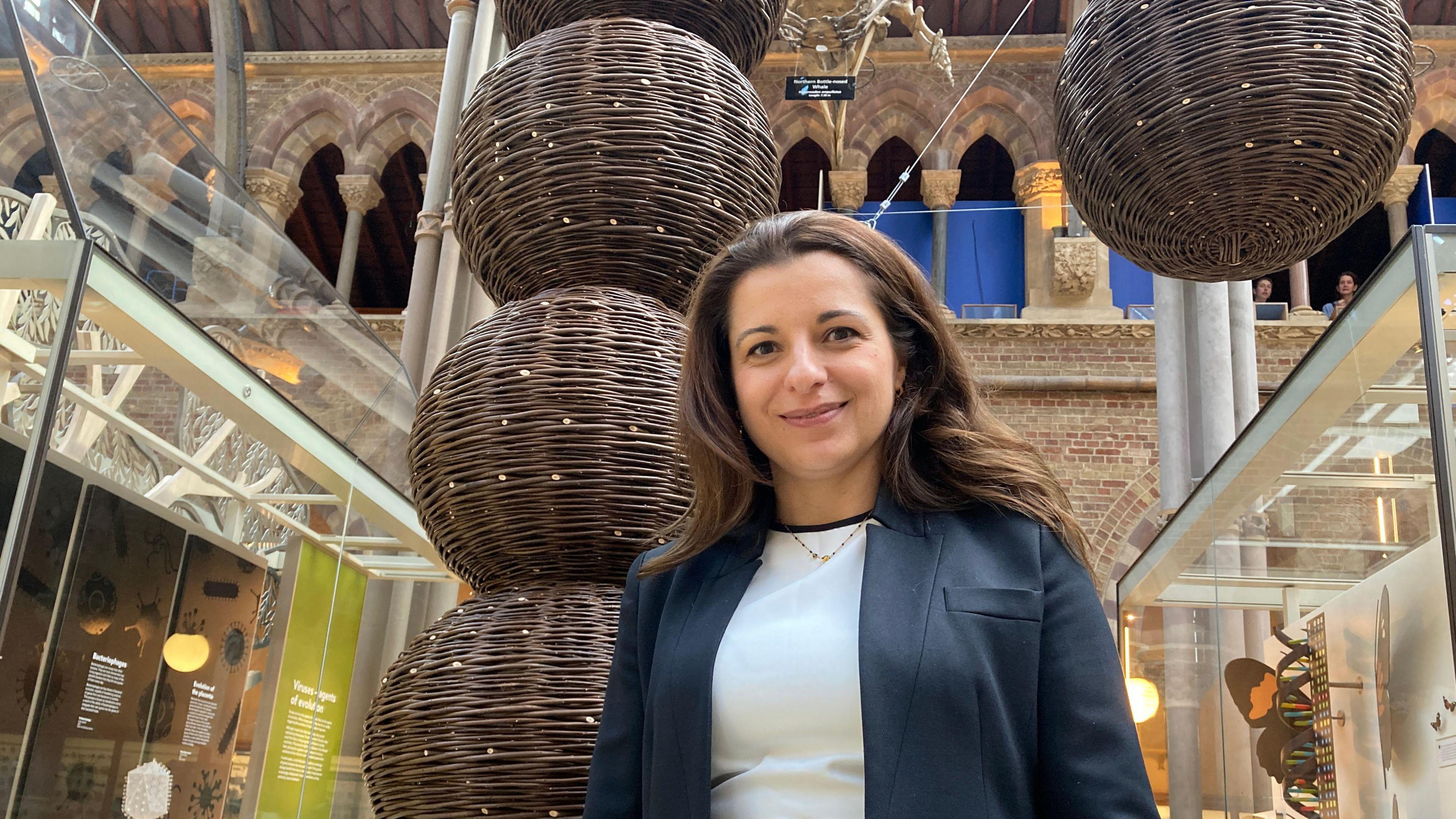 Prof Daniela Ferreira wearing a blazer, stood in front of three willow balls stacked on top of one another. They represent the pneumonia molecule.