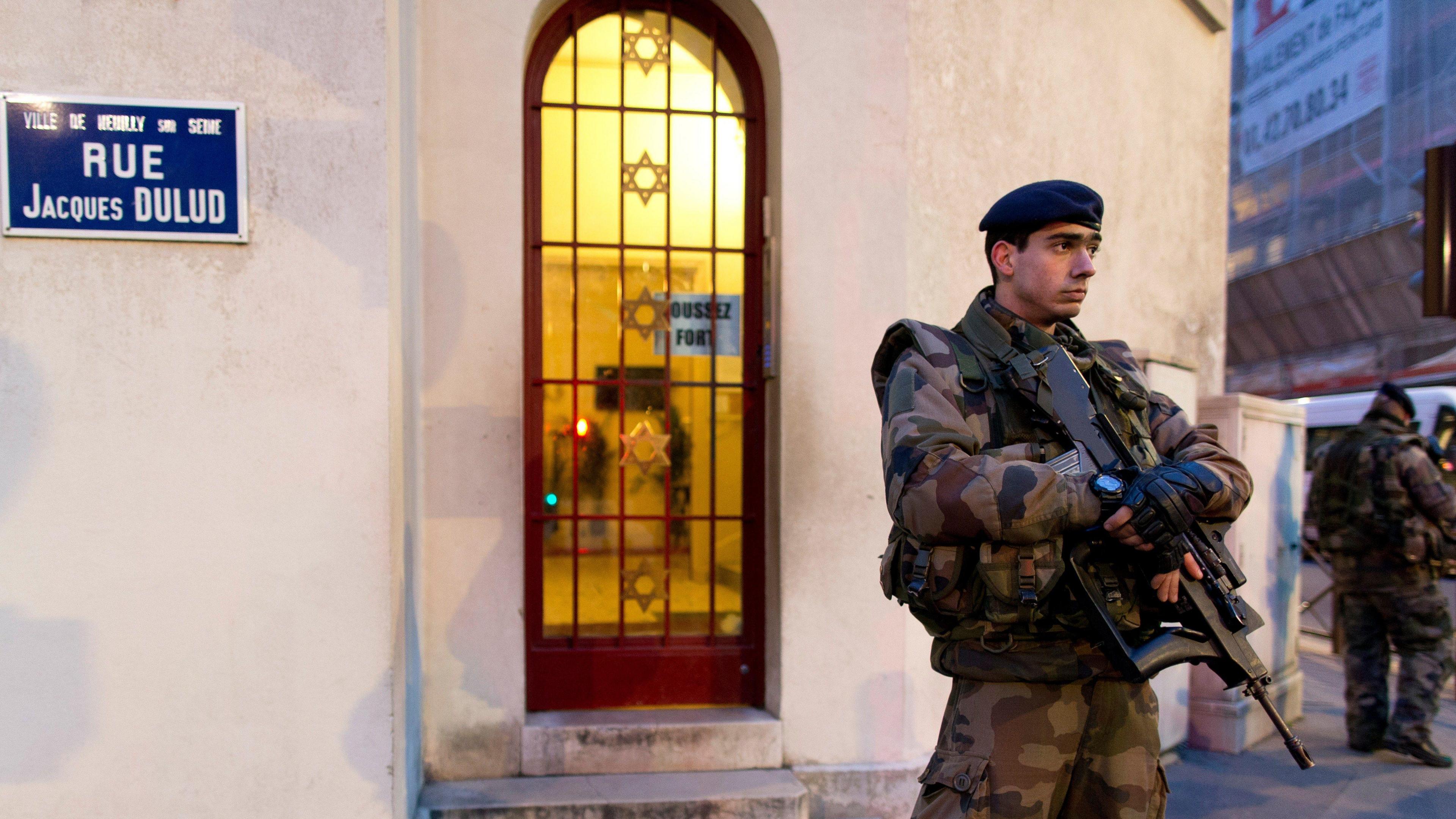 A French soldier standing guard outside a synagogue in a Paris suburb. He is dressed in military fatigues and is carrying a rifle.