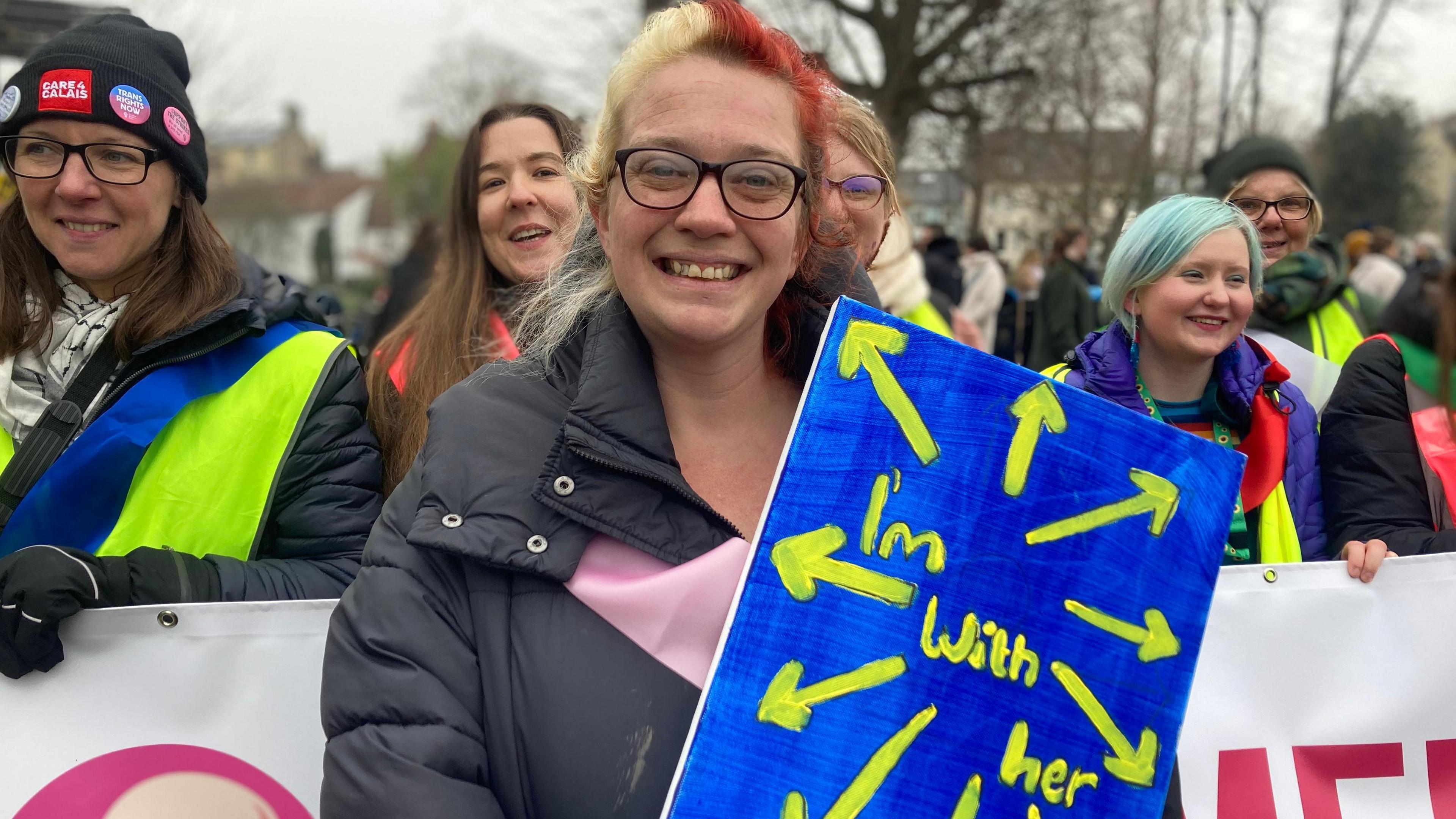 Alix Valentine, with half her hair dyed blonde and the other half auburn, wears a black quilted coat and holds a blue and yellow placard with arrows and the words: I'm with her. She is standing in front of female protesters holding a banner.