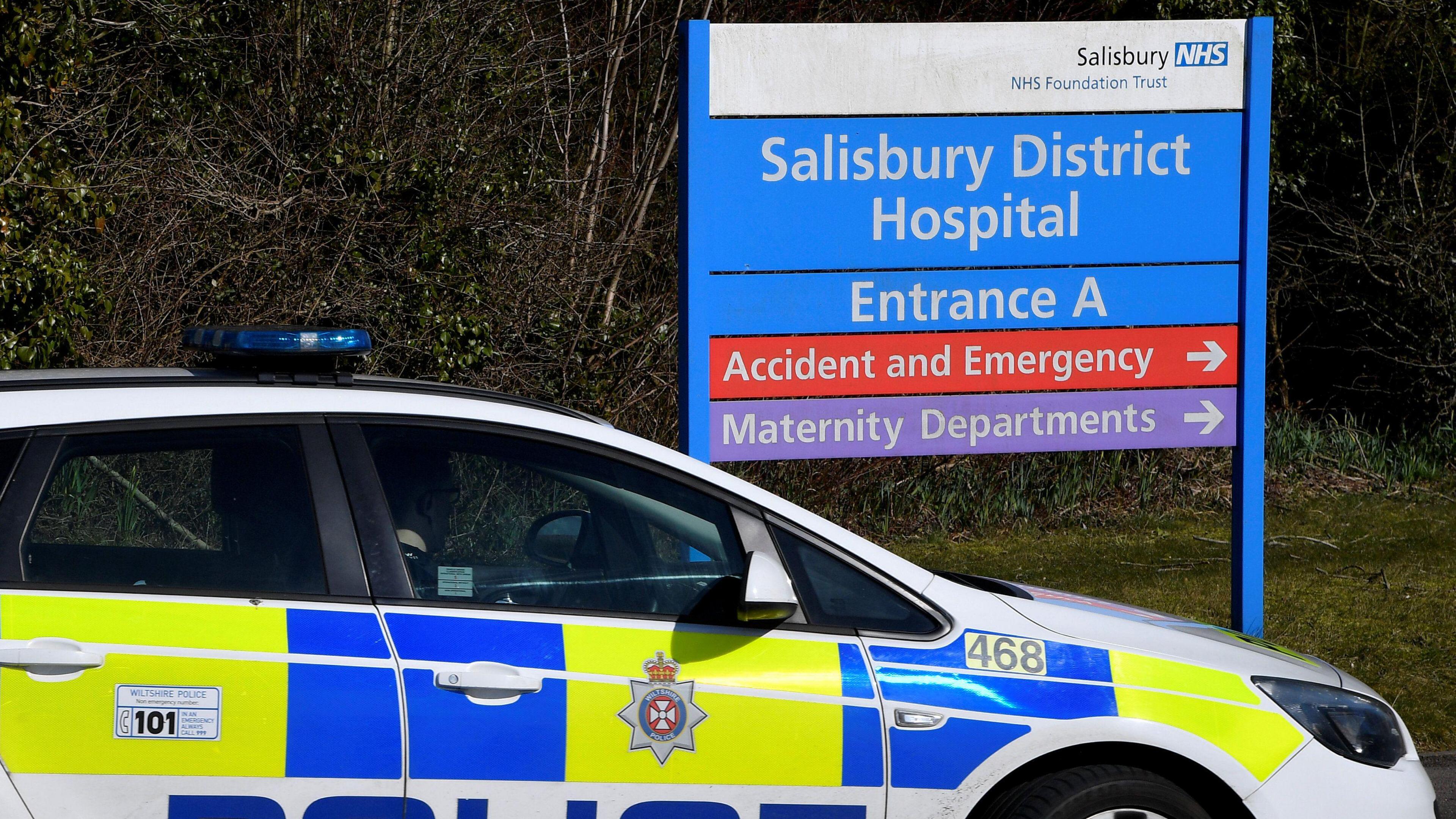 A police car, parked next to a large blue and white sign which says "Salisbury District Hospital". 