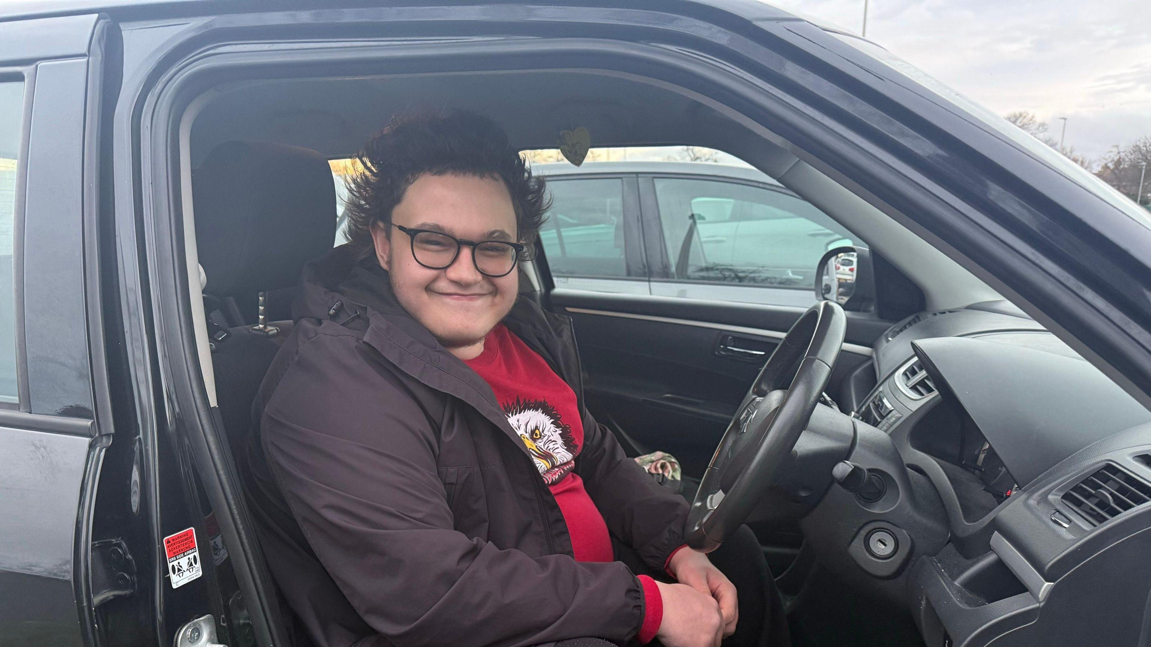 Sam Firbank sitting in the driving seat of his car. He is wearing glasses, a red T-shirt and a black jacket.