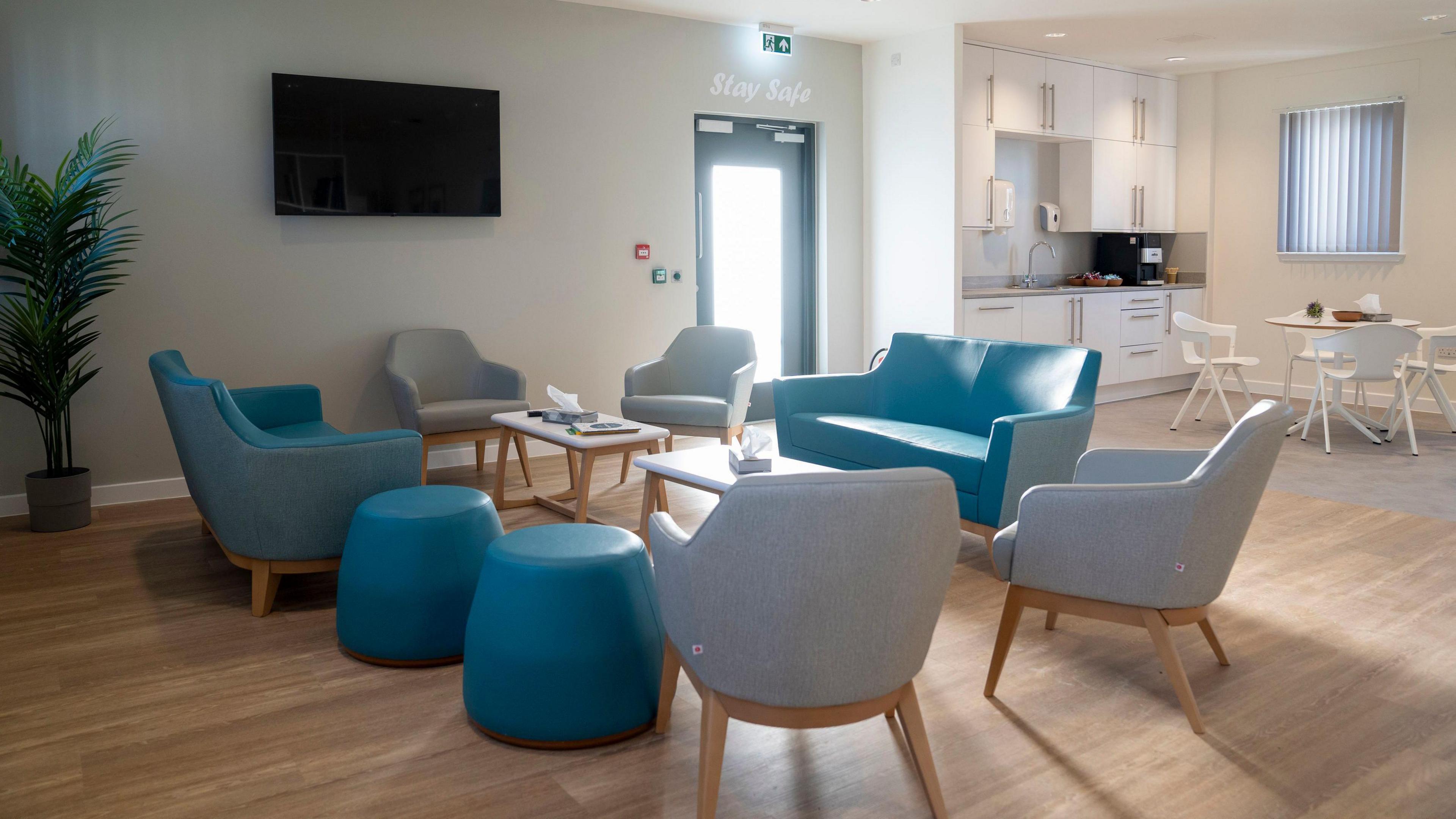A lounge area with blue and grey comfy chairs facing a table and a kitchen area in the background with a kitchen table. A TV is on the wall and the floors are wooden and the walls are painted a neutral grey/white colour