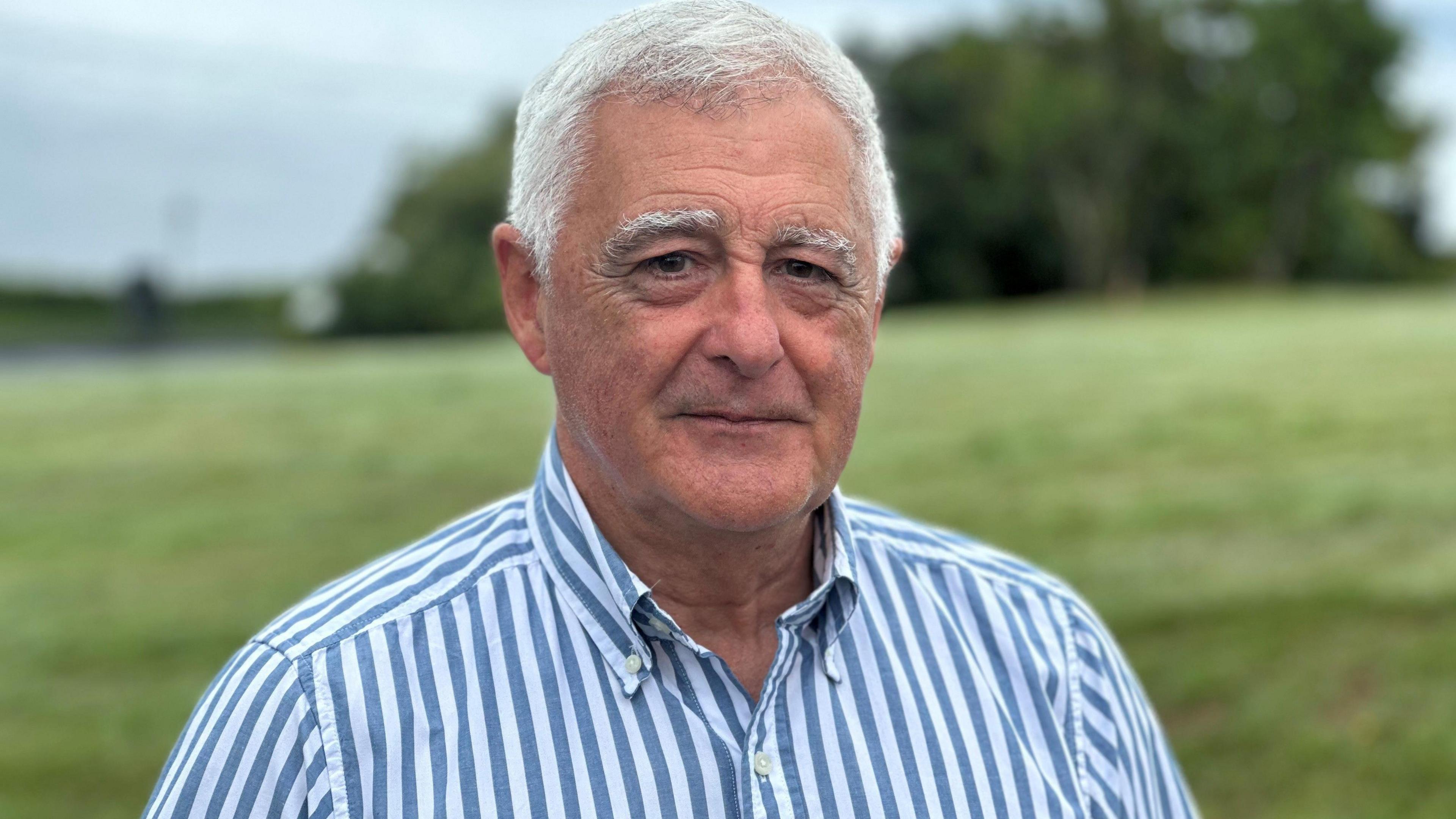 Charles Guard, who has short grey hair and is wearing a blue and white striped shirt. He is standing in front of a grassed bank.