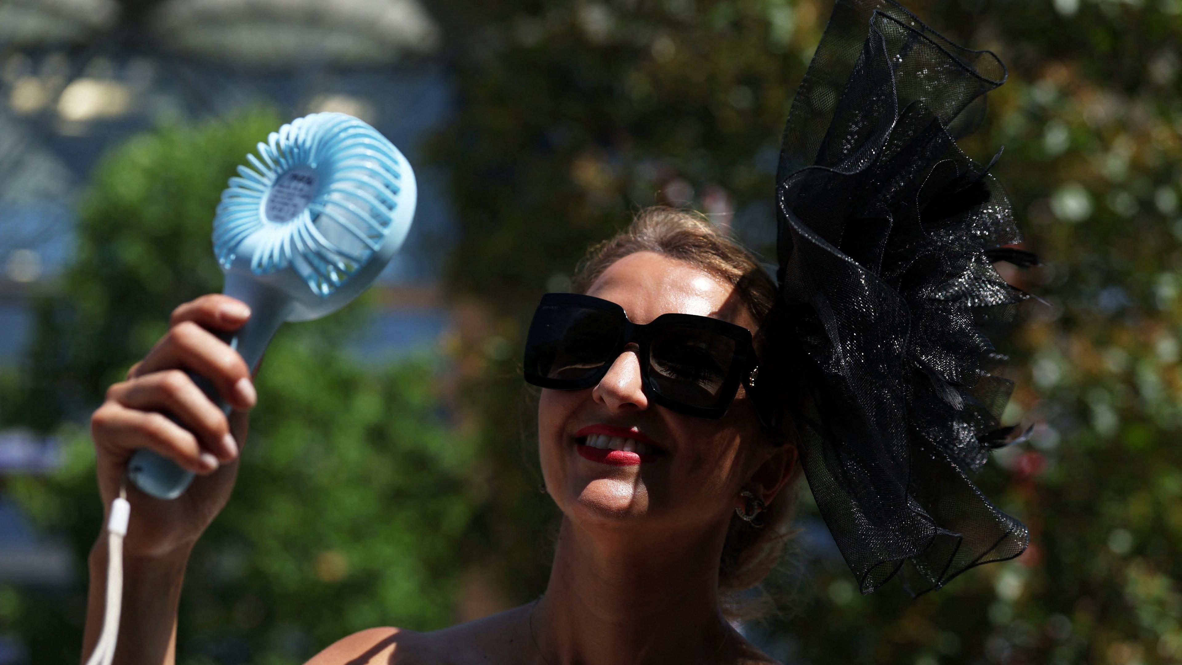 A woman is smiling and is standing holding a blue handheld fan in front of her face as she wears a black fascinator and black sunglasses. 