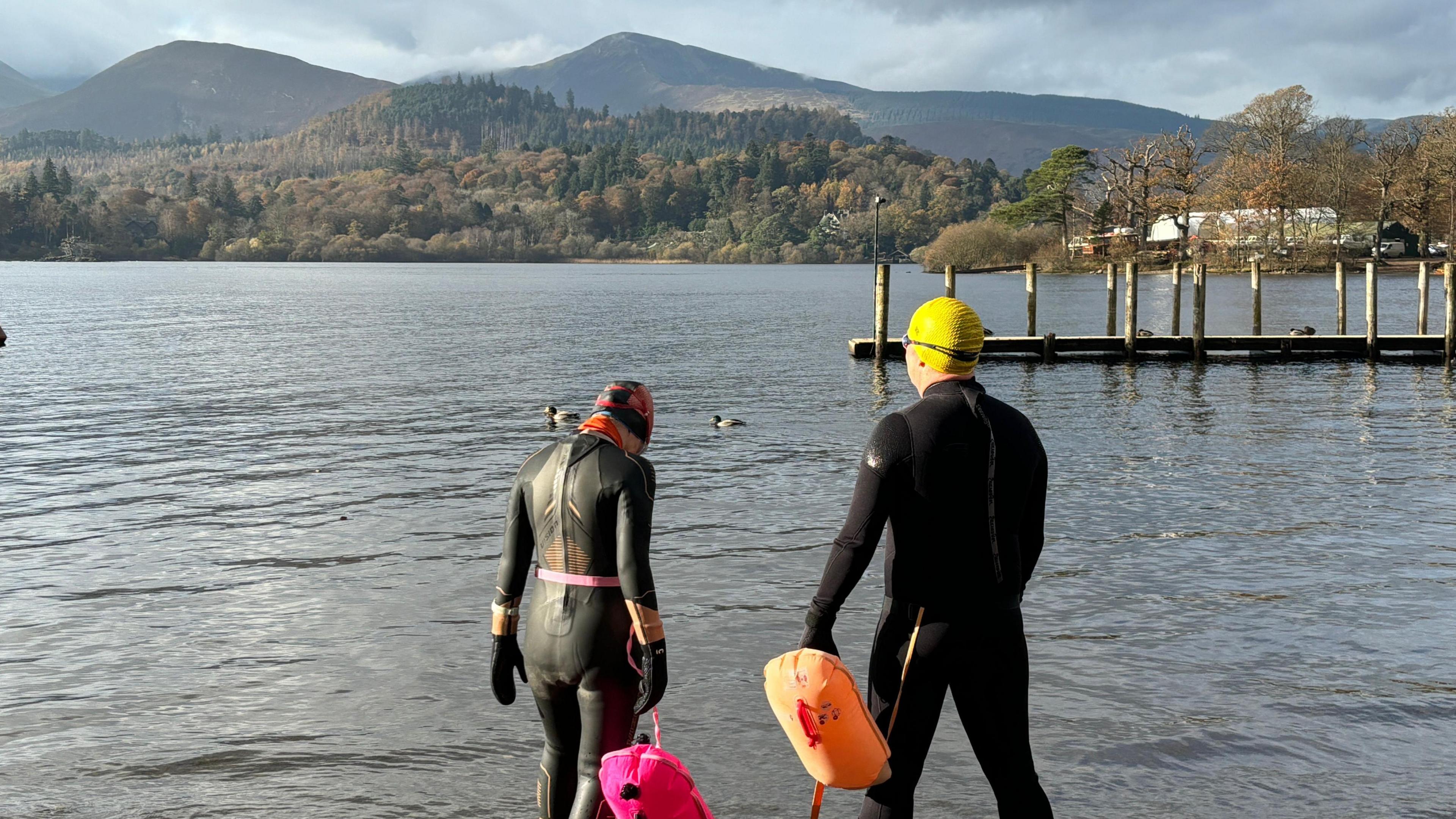 James Philips and Joanne McKenzie standing on the edge of the lake in their wetsuits. Mountains and forests can be seen in the background.