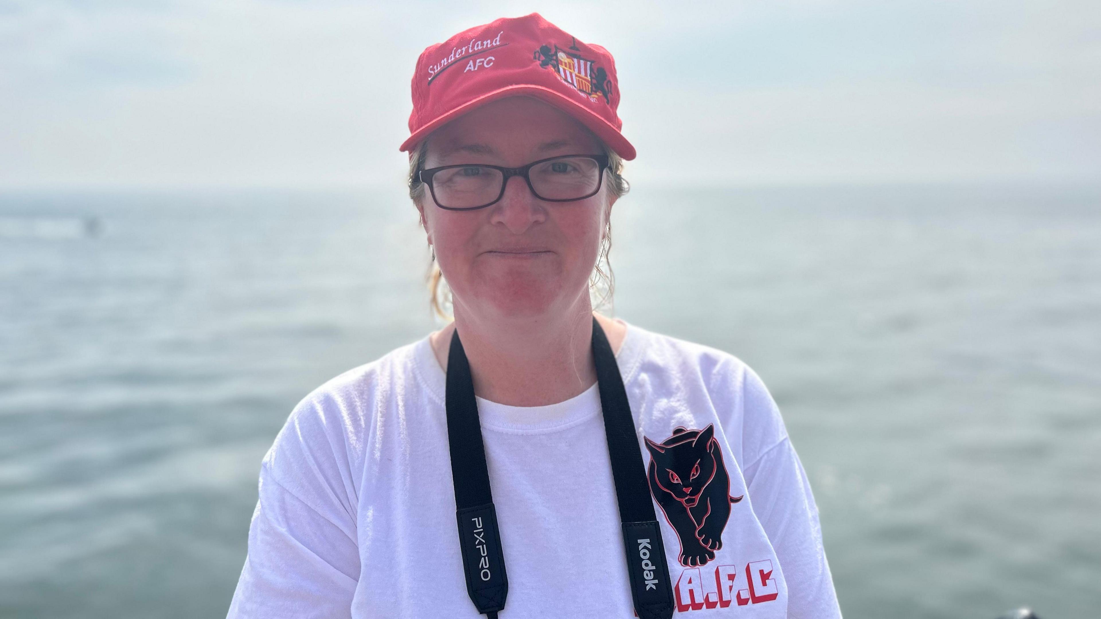 Kerrie Moorhead with black rimmed glasses, wears a red hat which says 'Sunderland AFC' on top, and a white T-shirt which says 'A.F.C' on the chest. She is looking at the camera and there is a black Kodak camera strap around her neck. The sea can be seen behind her.