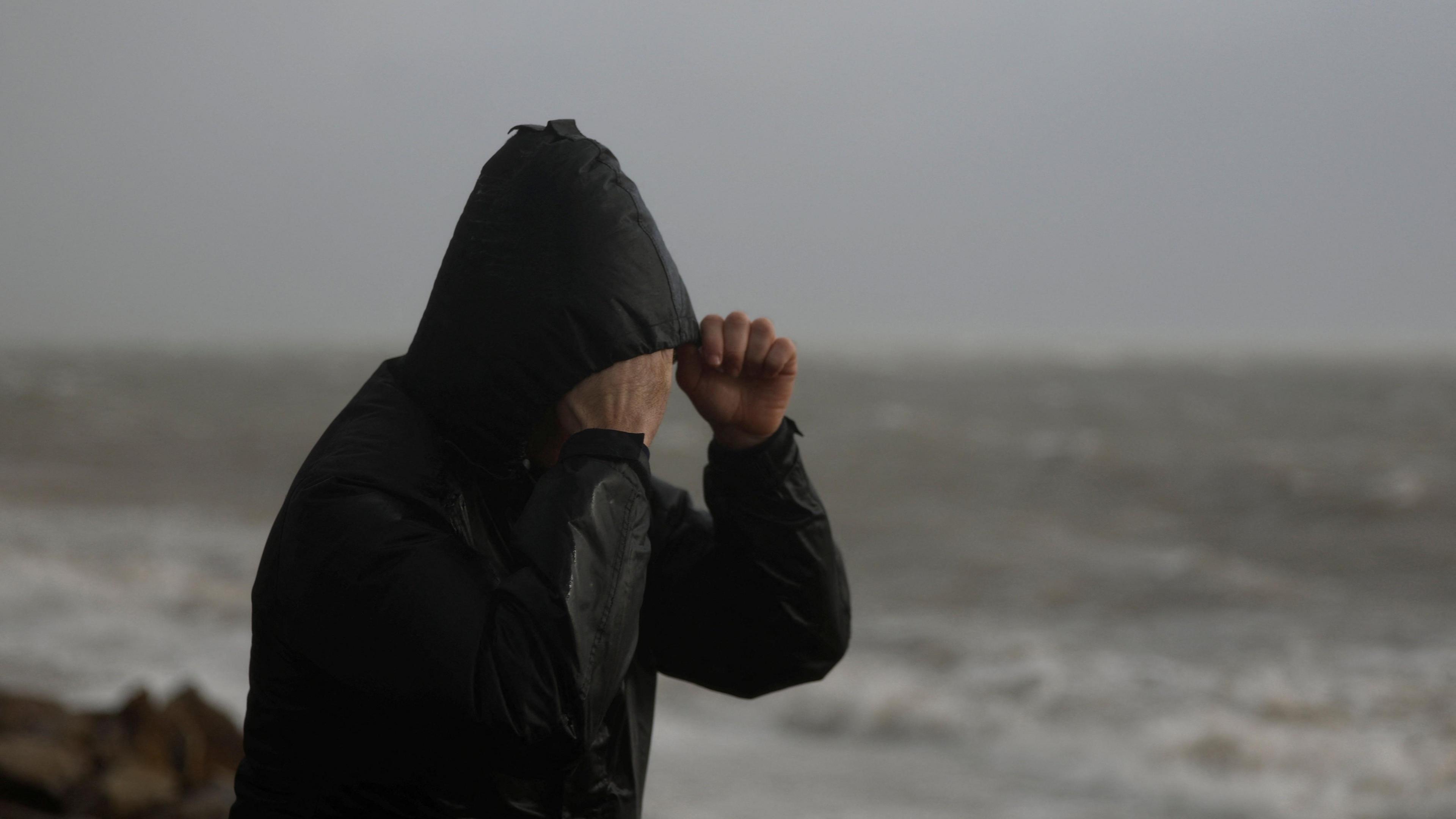 A man wearing a black hooded raincoat trying to protect his face from the rain and tide - pictured in the background.