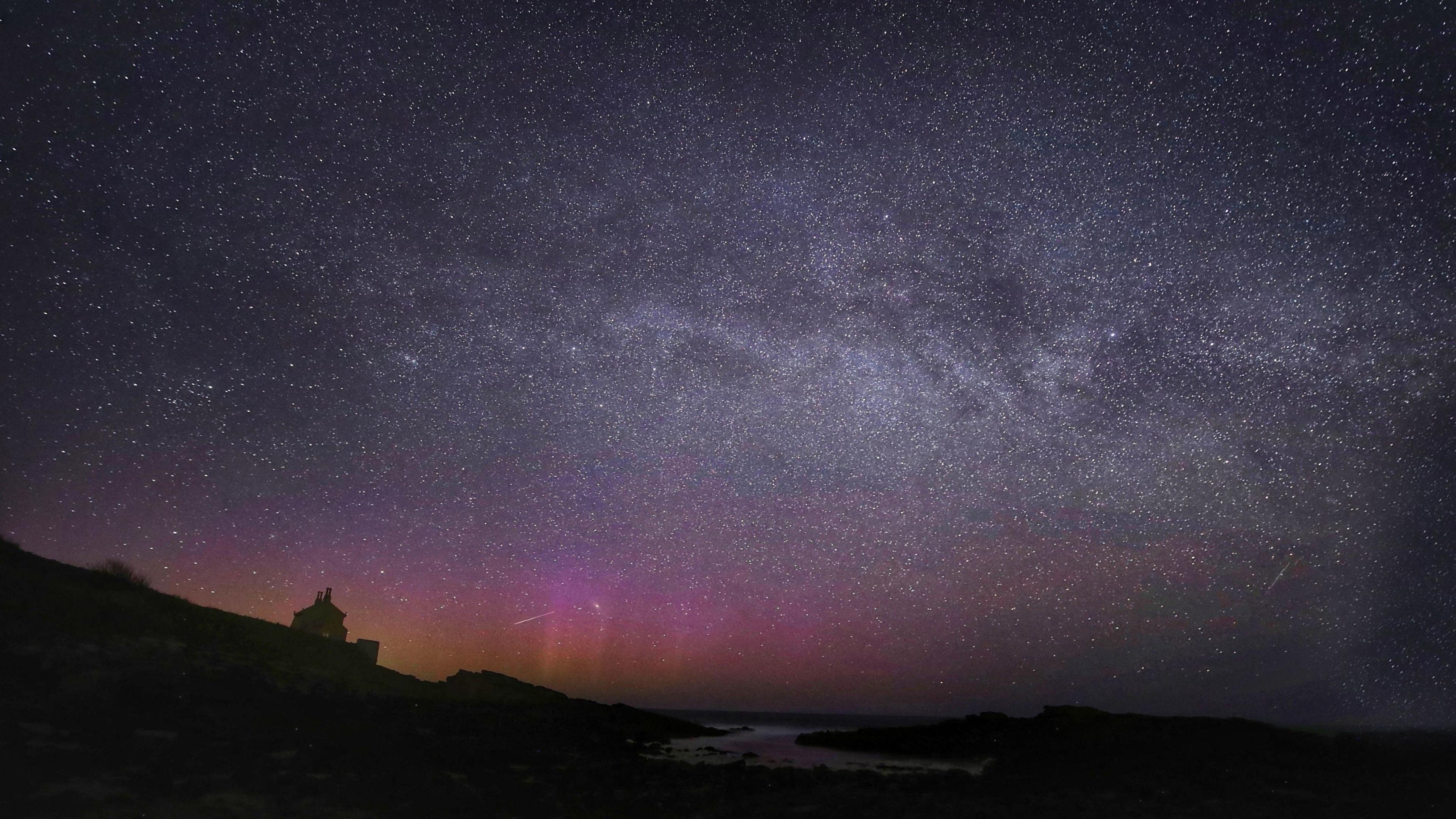 A blue and purple sky showing pinks and oranges of the Northern Lights, a silver colour swirl of the Milky Way and white streaks of Lyrid meteors falling through the sky with dark outline of a hours and coastline