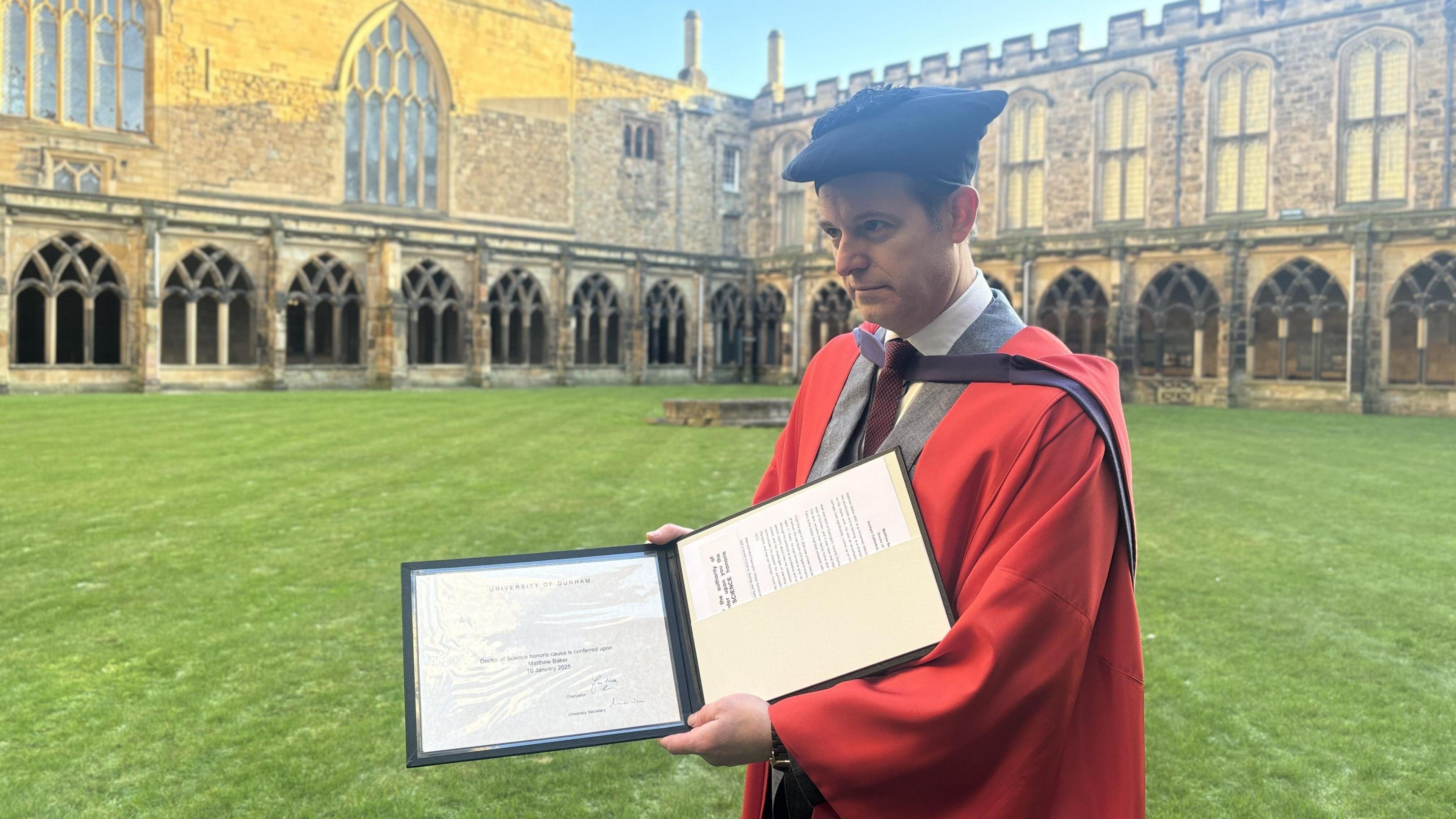 Matt Baker wearing a red graduation cloak, on top of a grey blazer with a maroon tie, holding up a two sided folder which holds his certificate and documents. 
