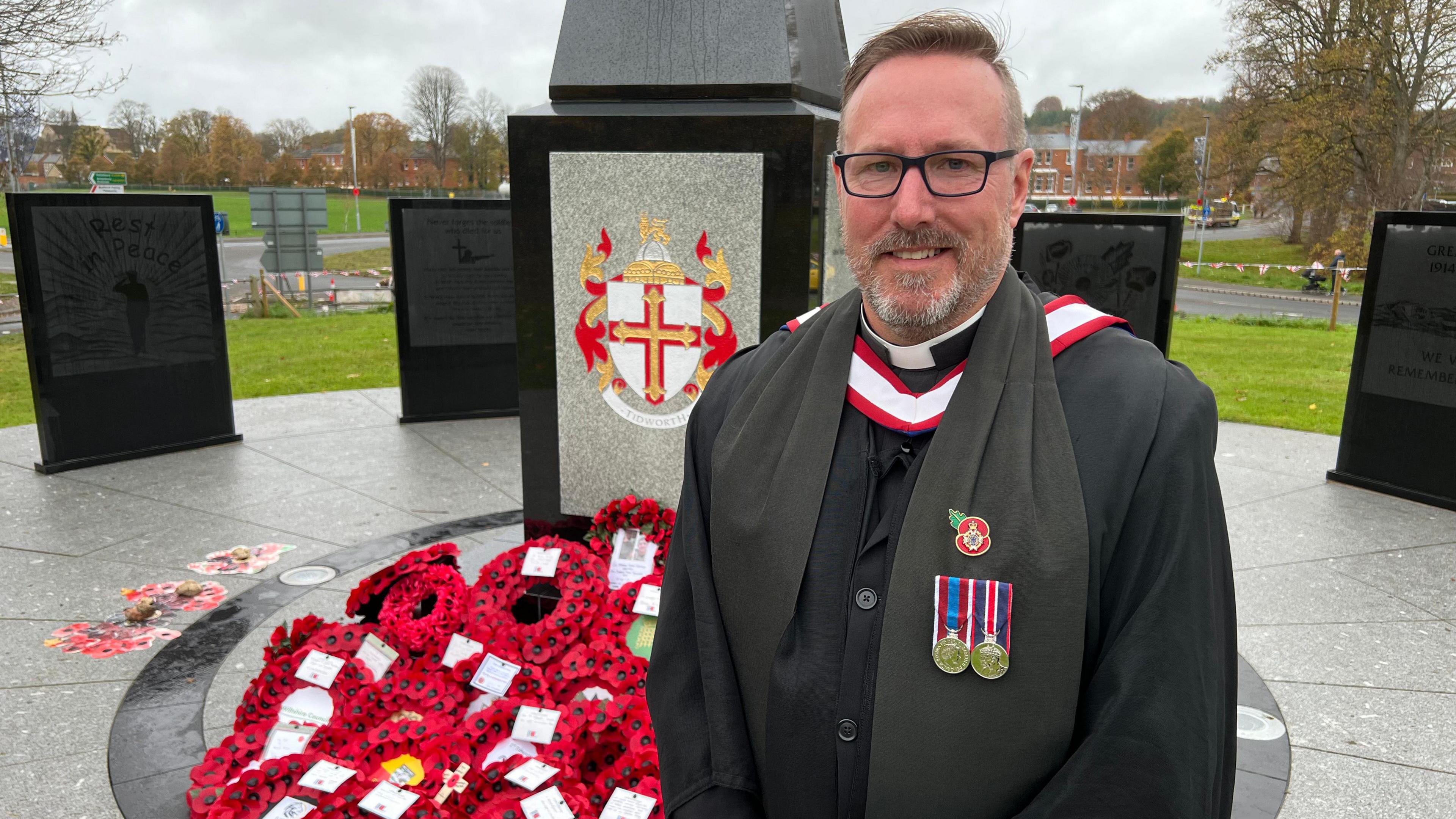 Rev Maj Gary Birch. He is dressed in black and has a poppy badge and two medals on his chest. A war memorial is behind him covered in poppy wreaths. Mr Birch is looking at the camera and smiling.