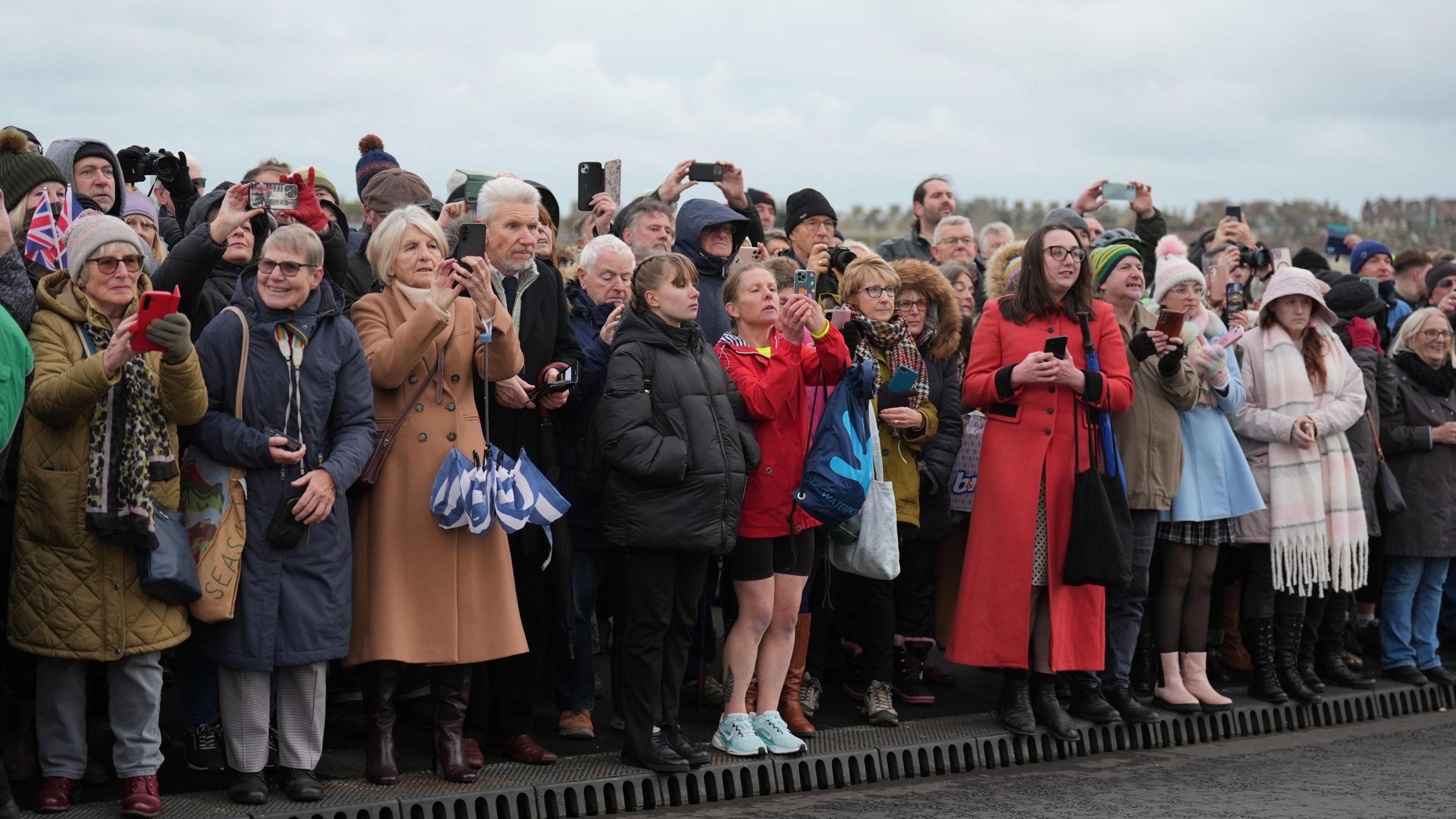 A crowd of people gathered on the side of the road that creates the Gull Wing Bridge. Many have their phones out trying to take pictures of Princess Anne, who is behind the camera.