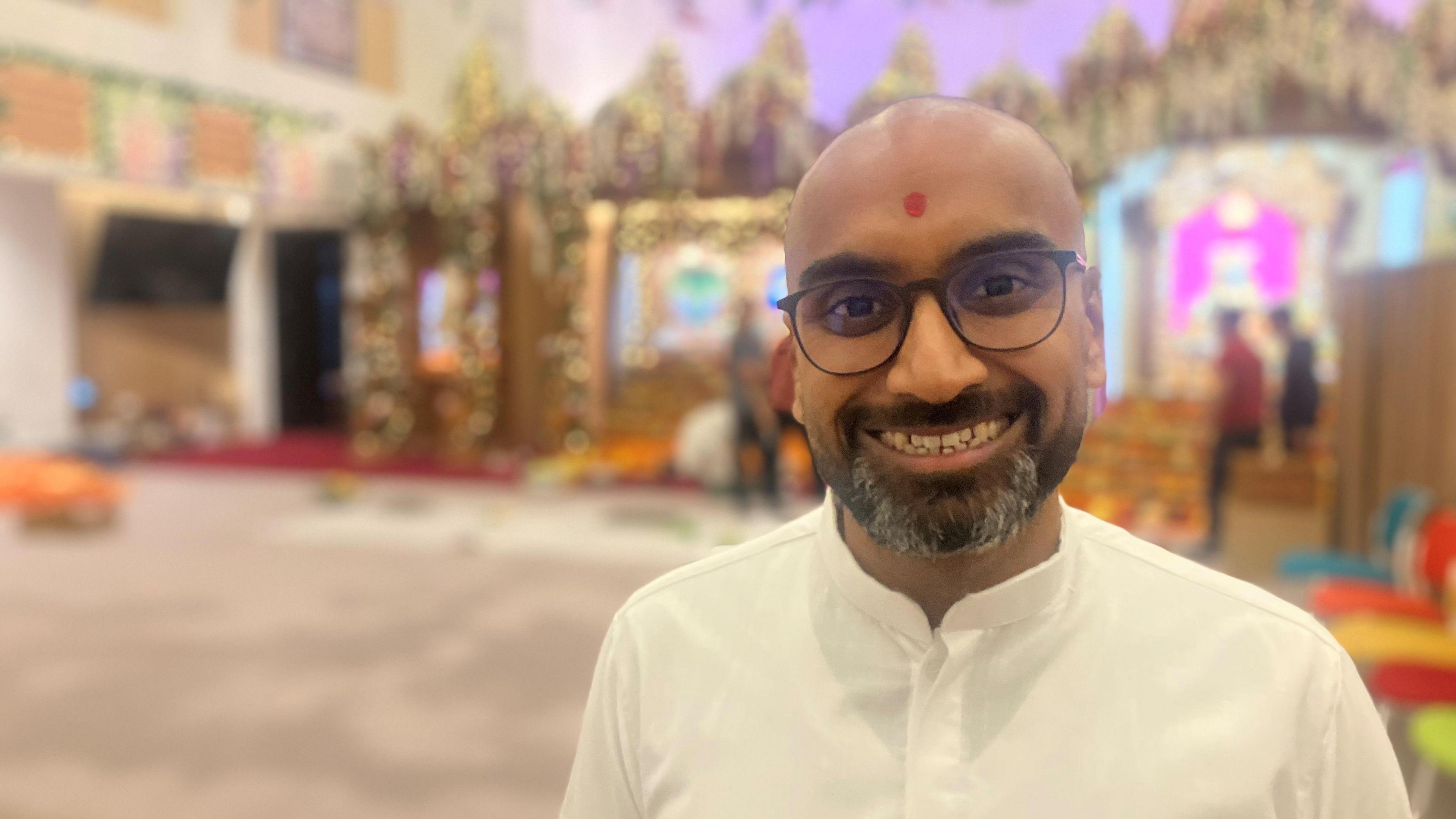 A man stands inside a colourful temple wearing a white button up shirt. He has a red mark painted on his forehead and is smiling.