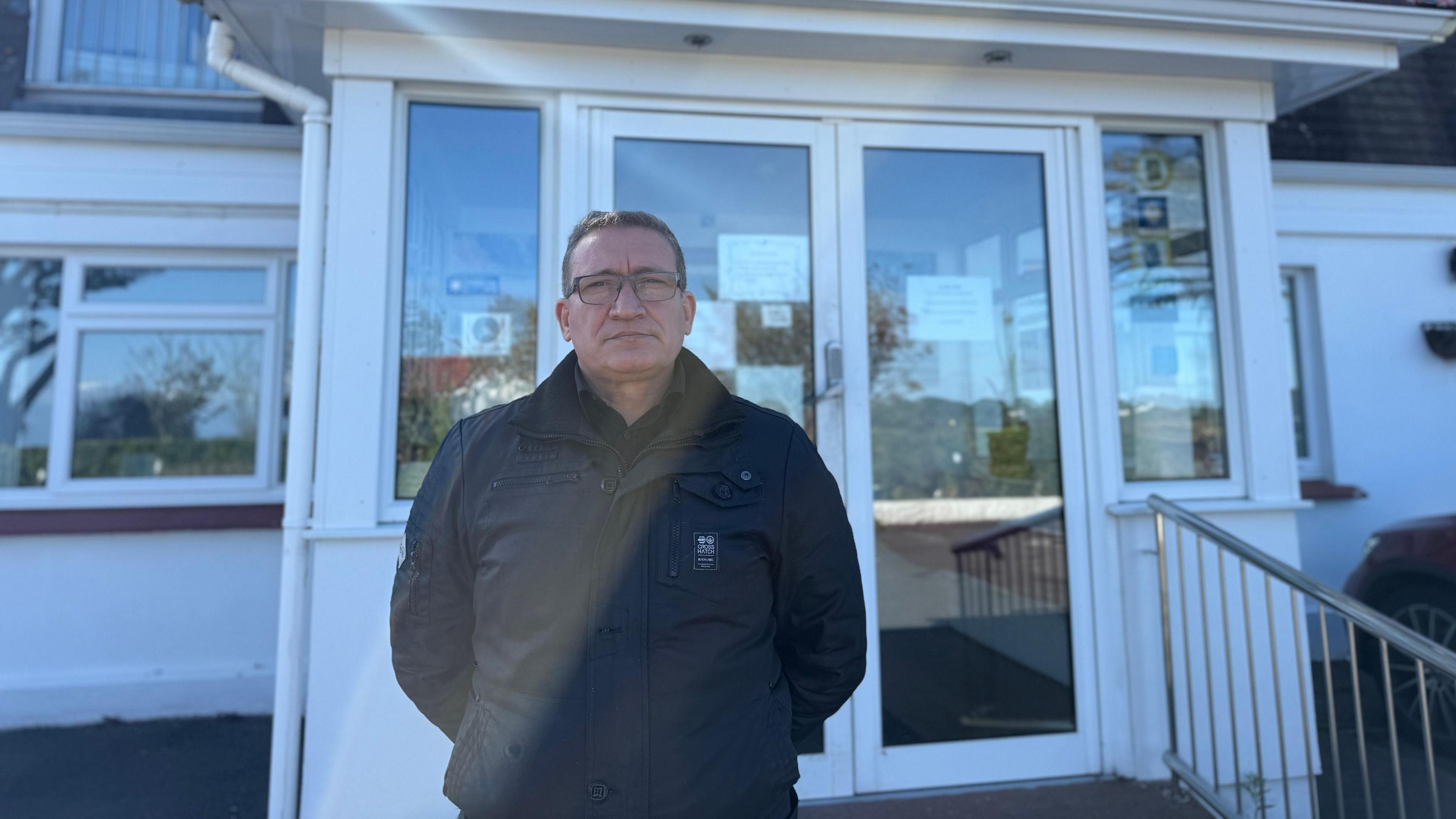 Image shows Mr Ashraf Elsergany infront of the white doors of his hotel. He wears a black jacket, with glasses and short brown hair.