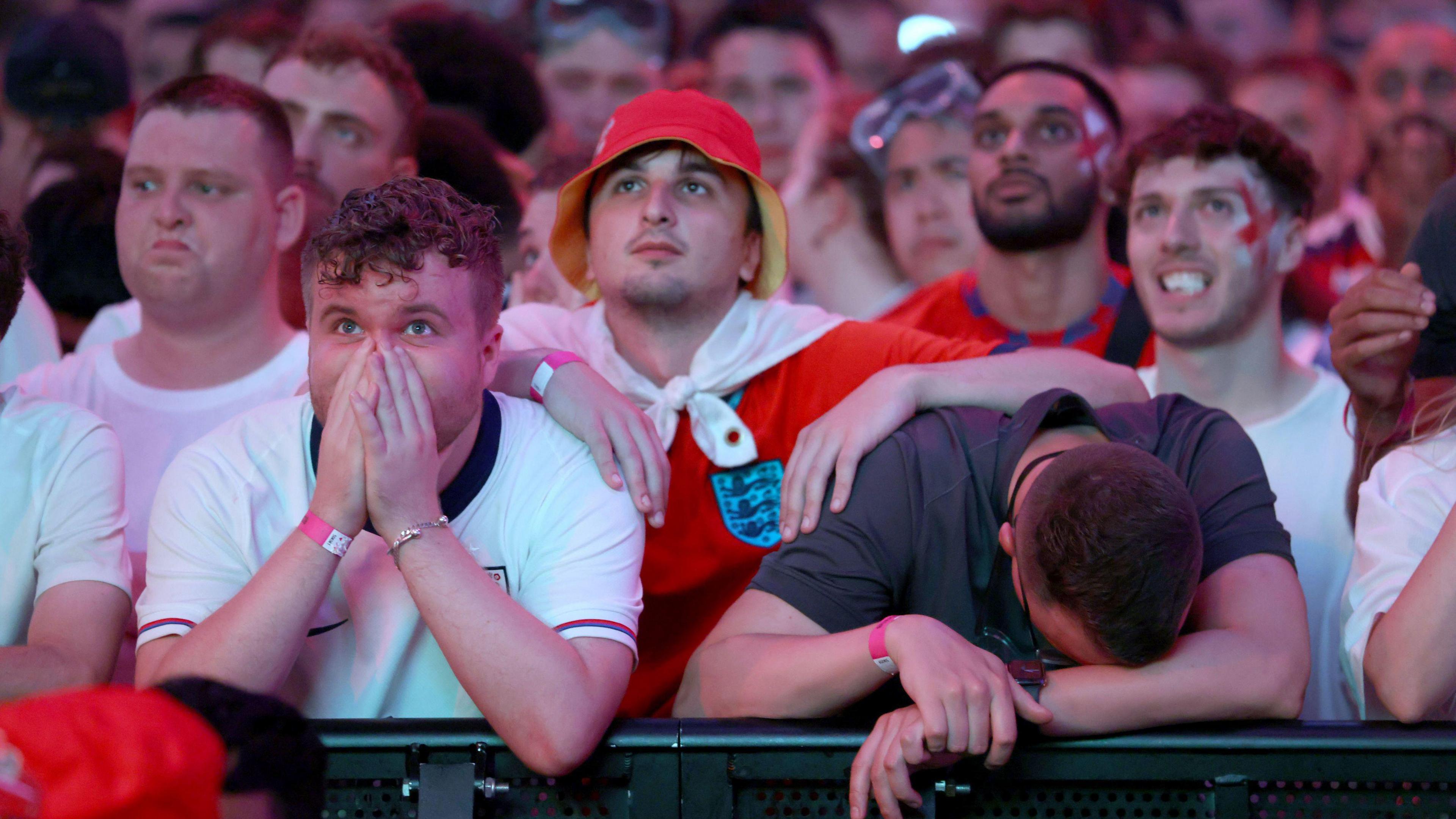 Men in red and white England shirts with a disappointed look on their face as England lose the Euro 2024 final to Spain. One man in a grey T shirt rests his head on his arm. 