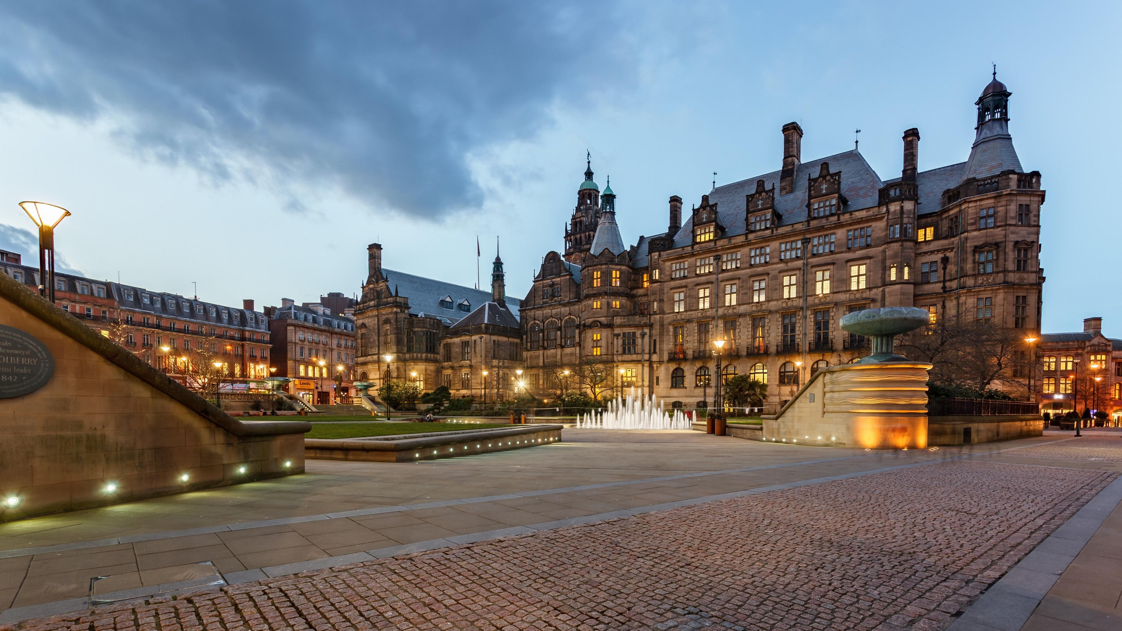 Sheffield Town Hall photographed at dusk. A large stone-built building stands behind a paved area with a large fountain and grassy areas. 