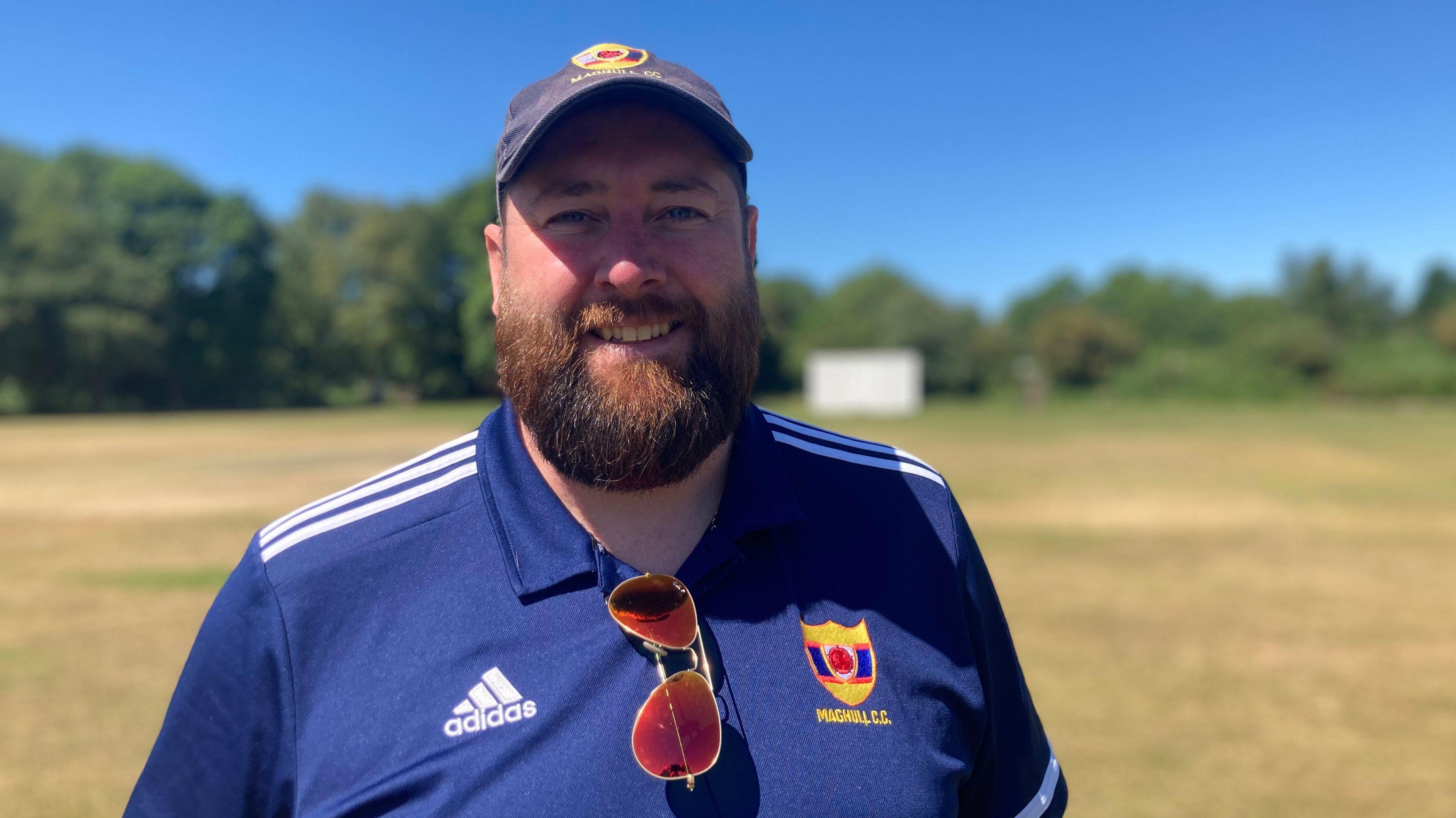 Maghull Cricket Club chairman Adam Lloyd smiles while stood on the club's cricket ground. He has a big beard and is wearing a cap and club t-shirt.