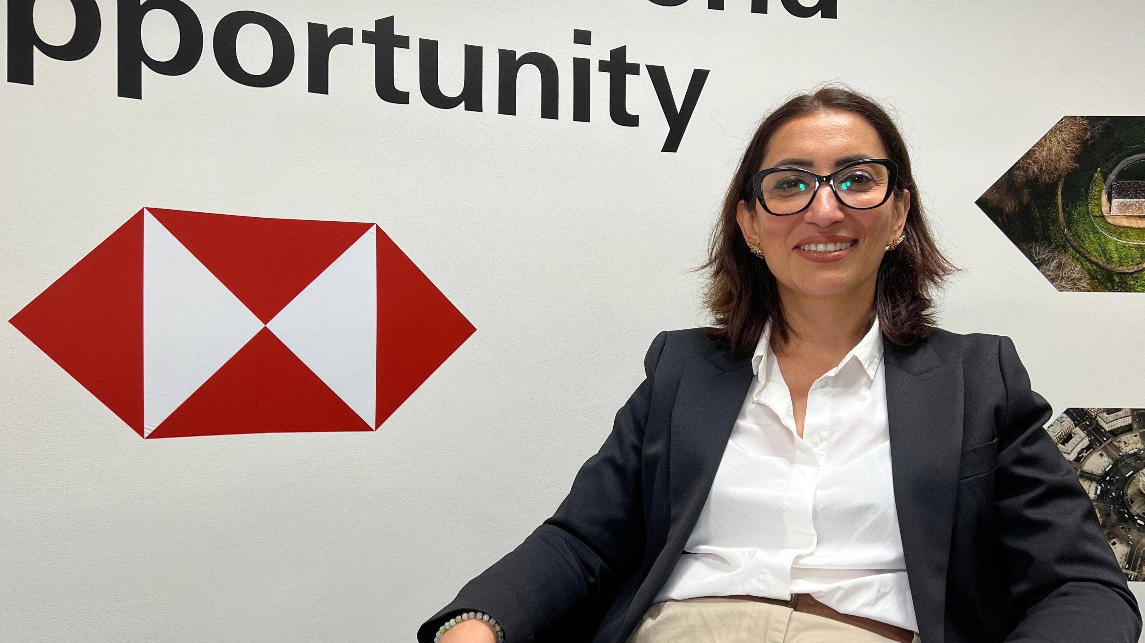 Elvina is seated on a chair holding a piece of paper. Behind them is the HSBC logo and the slogan 'Opening up a world of opportunity'