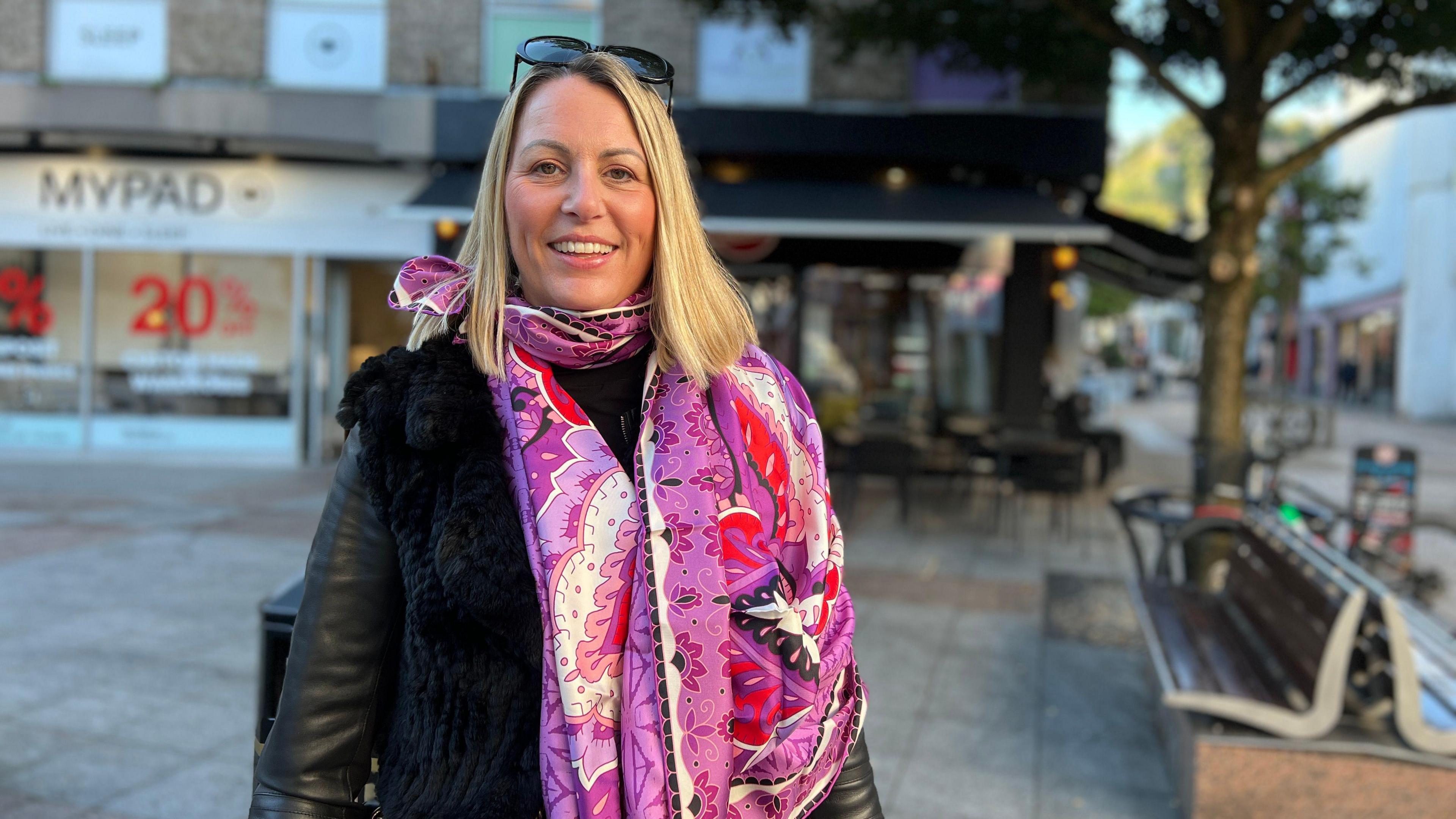 Bex smiles at the camera and she's wearing a black, leather jacket and has a purple long scarf with floral decorations on it. She has blonde hair and her sunglasses are on the top of her head. There's a bench on a concrete block to the left of her and a shopping street in the background.