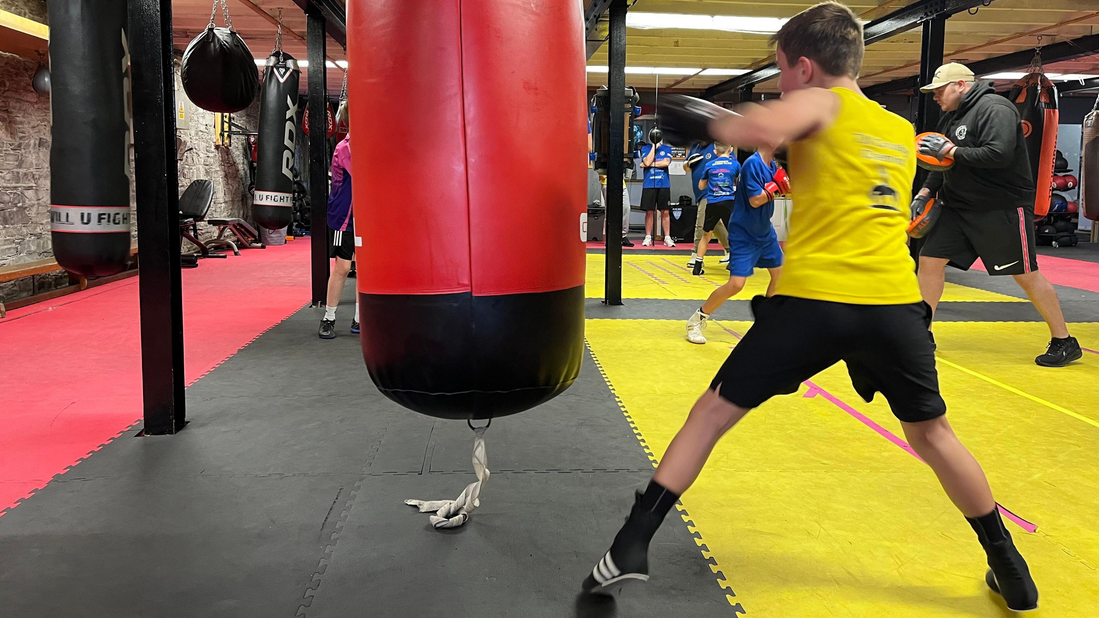 A boy in a yellow vest, black shorts and black boots punches a red punchbag.