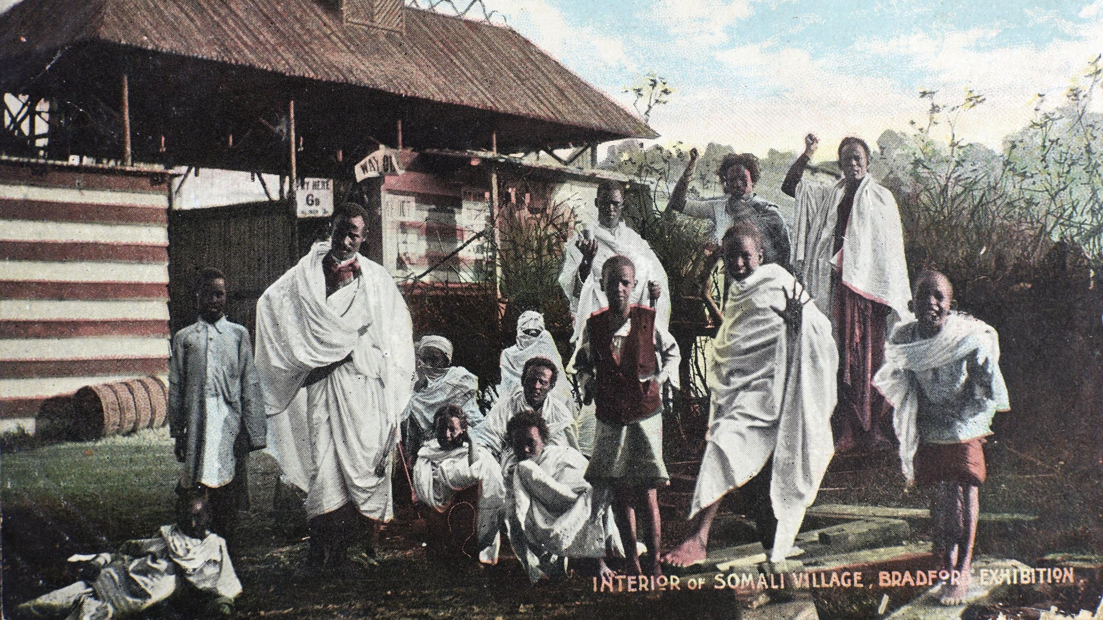 A group of men, women and children wearing white wrap style clothes in a traditional African style pictured in front of a large wooden hut. They are smiling and posing happily. The photo looks faded and old. In the right hand bottom corner are the words 'Interior of Somali Village, Bradford Exhibition'.