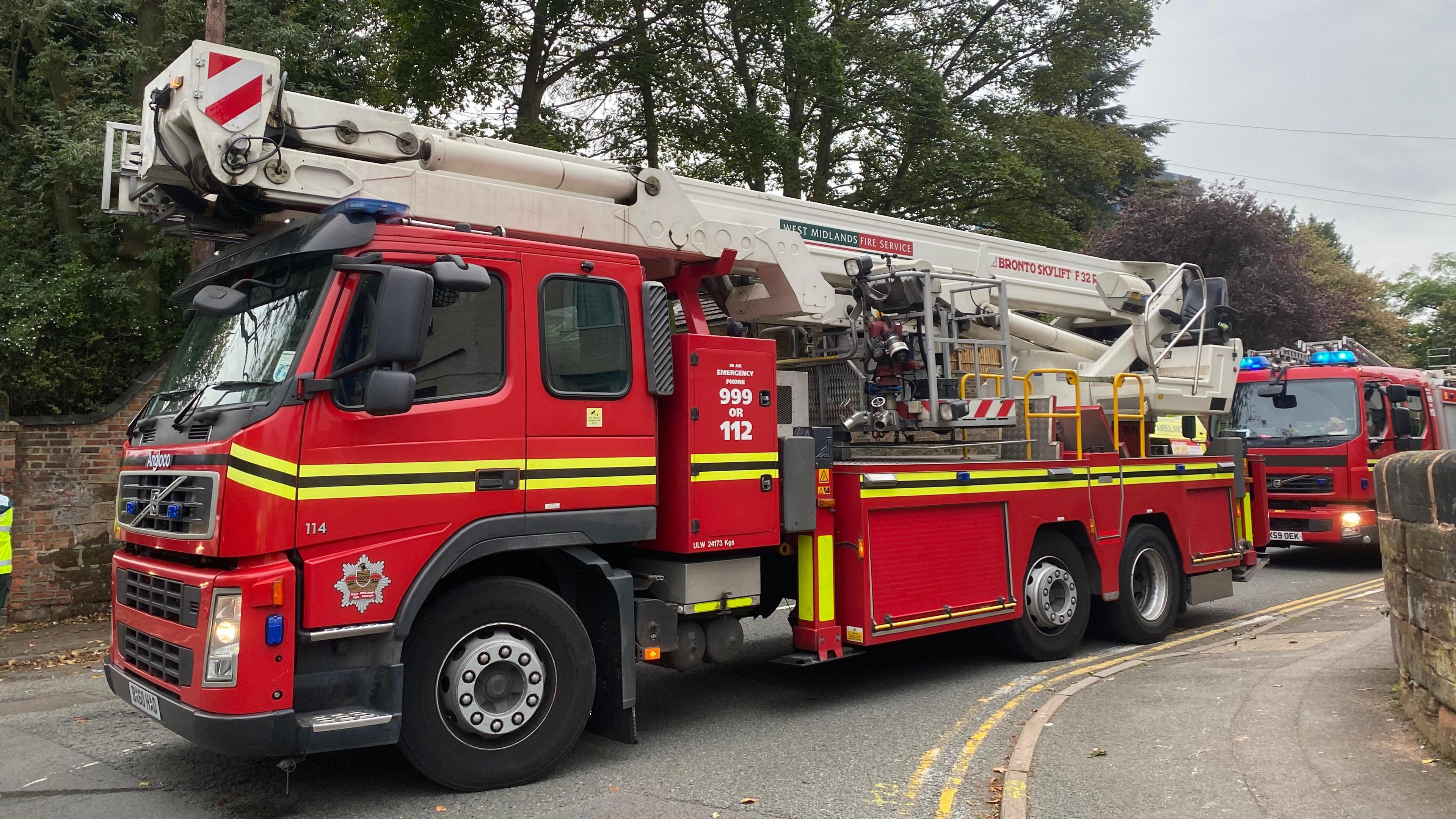 A stock image of a West Midlands Fire Service fire engine attending a different incident. The vehicle is parked on a road with another fire engine behind it. 
