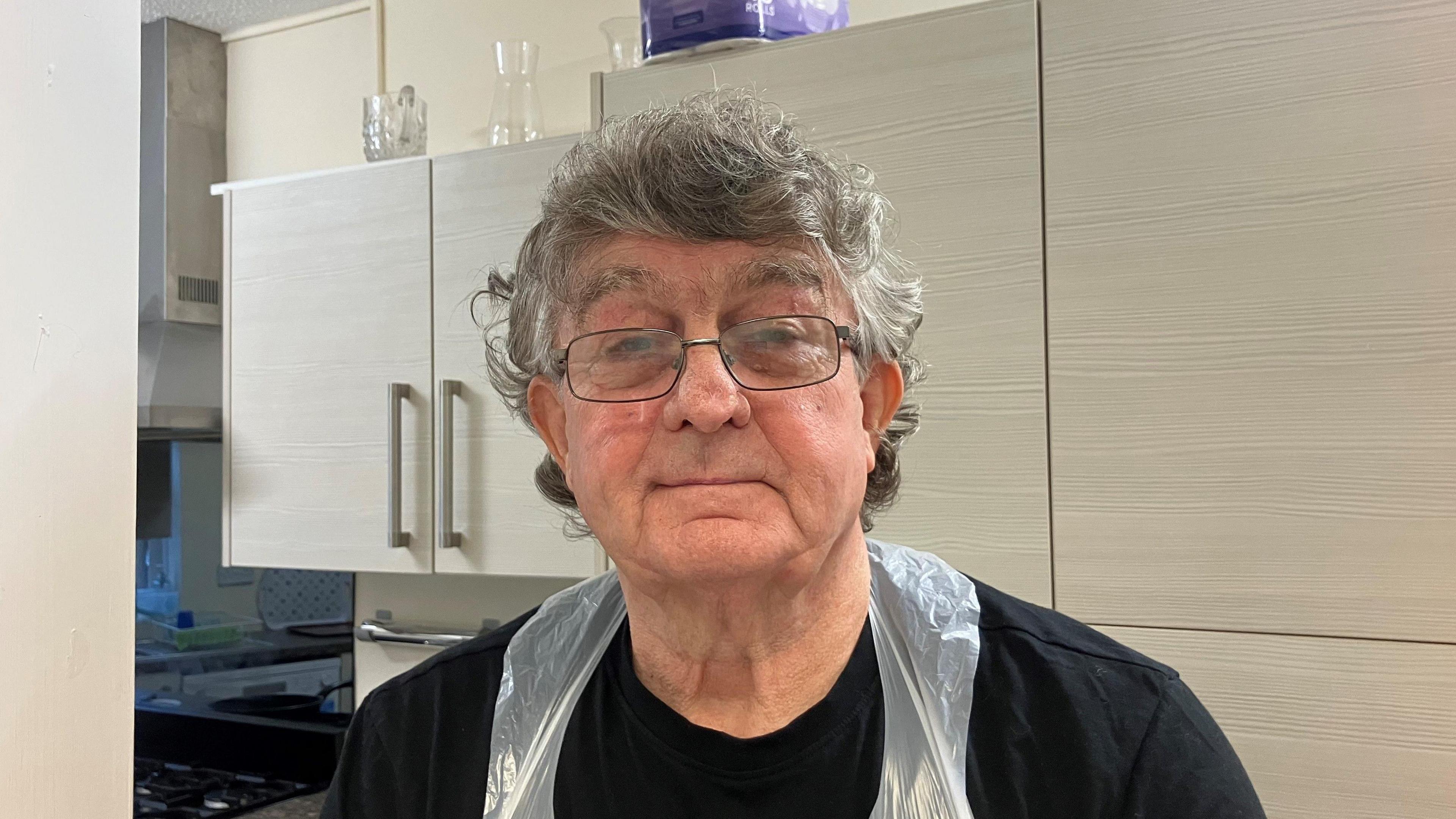 An older man with short grey hair and glasses. He is wearing a black t-shirt and a plastic apron and standing in a kitchen.